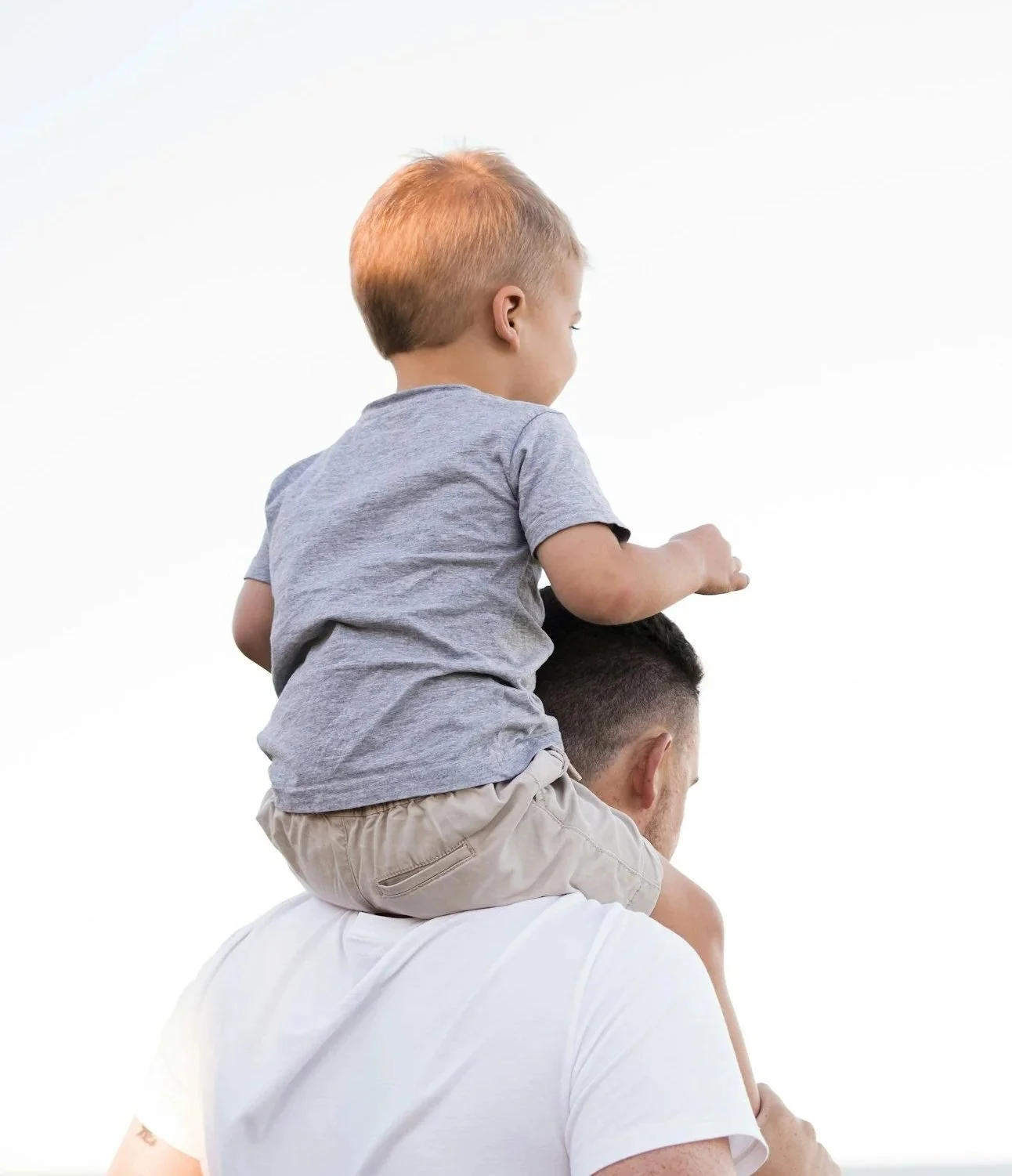 A child with red hair sitting on an adult's shoulders, wearing a gray t-shirt and beige shorts, against a white background.