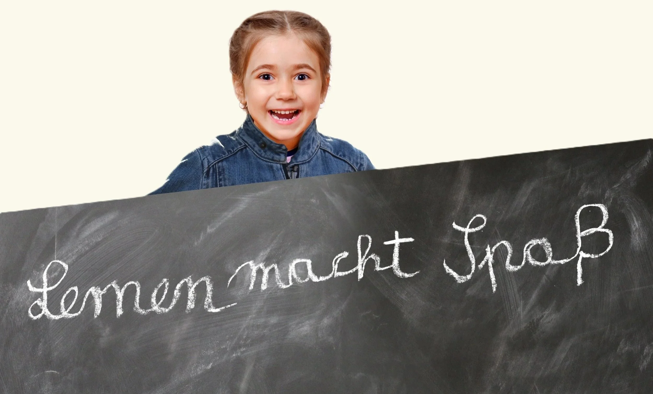 A smiling young girl with brown hair tied back, wearing a denim jacket, standing behind a black chalkboard with white chalk writing that reads 'Lemon', 'mash', 't &'. The background is plain and light-colored.