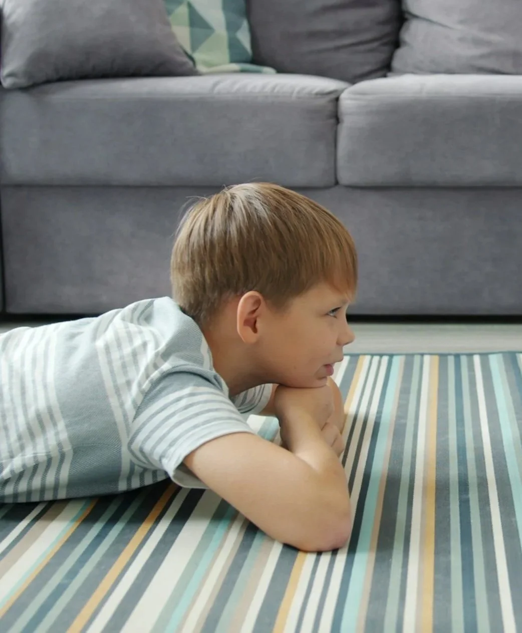 A young boy with red hair lying on his stomach on a striped rug, propped up on his elbows, looking intently at something off-camera in a living room.