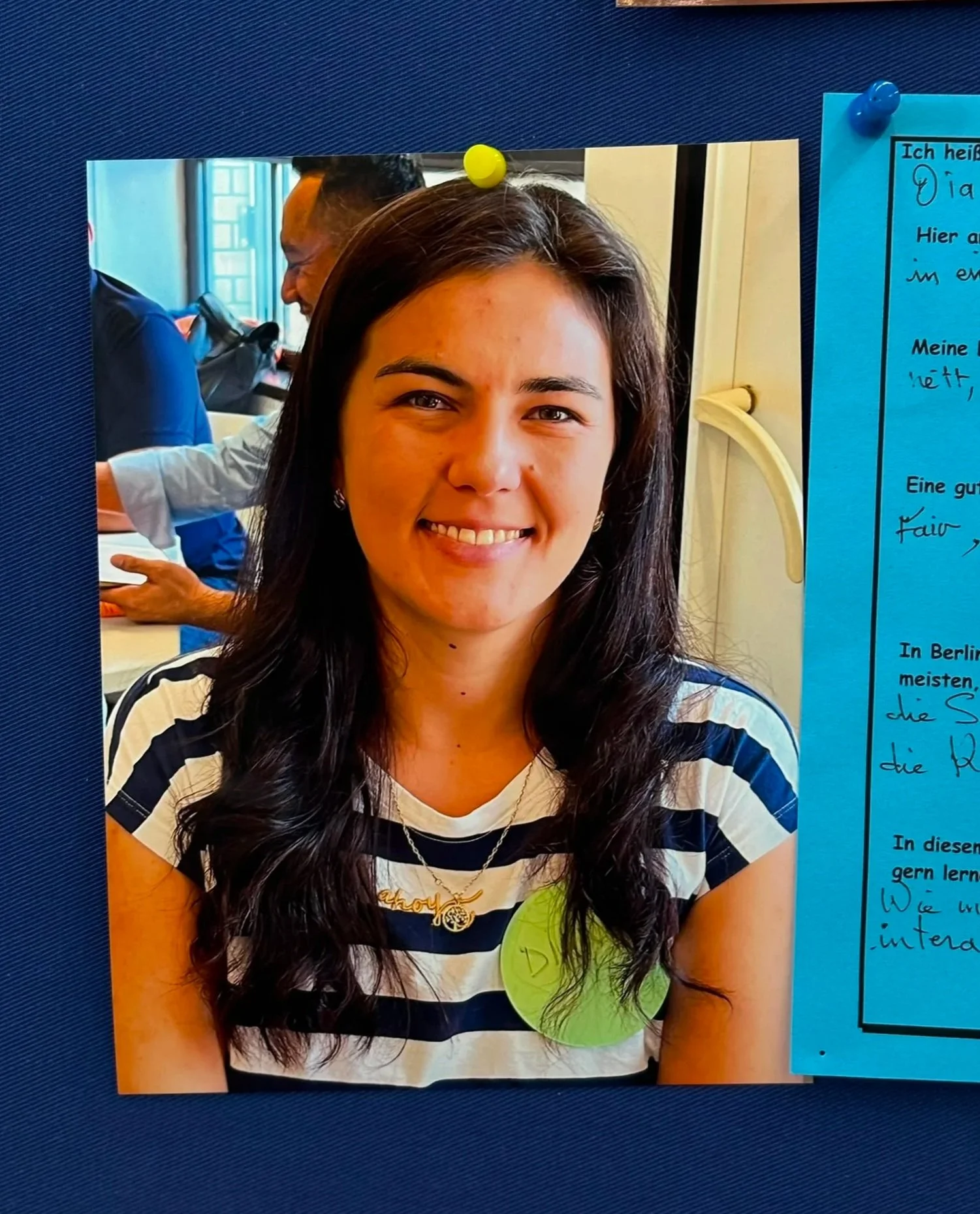 Photograph of a woman with long dark hair smiling, wearing a striped shirt and a silver necklace, with a person in the background. The photo is pinned to a blue textured surface with yellow and blue push pins, next to a blue paper with handwritten German text.