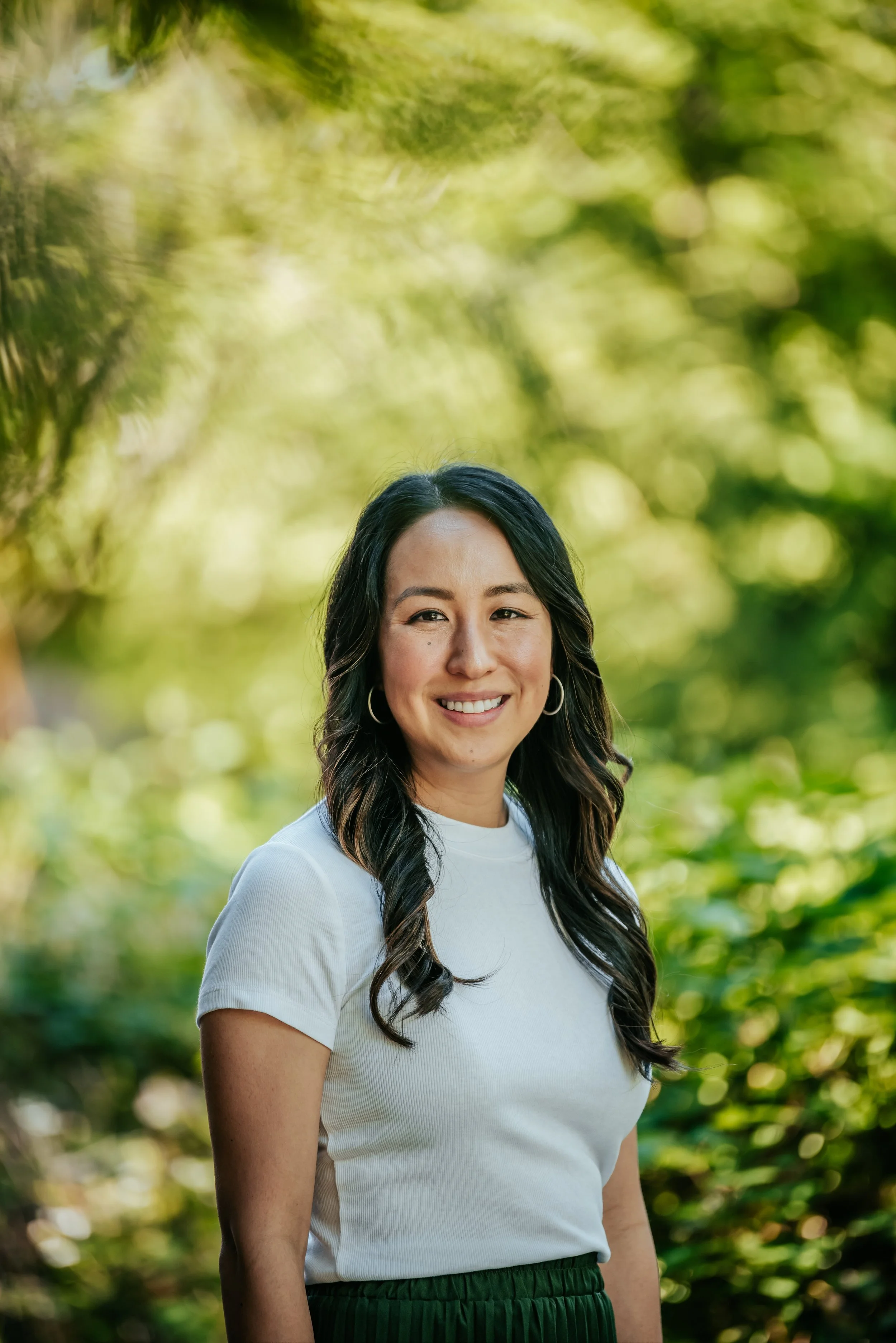 A woman with long dark hair, wearing hoop earrings, a white t-shirt, and dark green pants, smiling outdoors with a green leafy background.