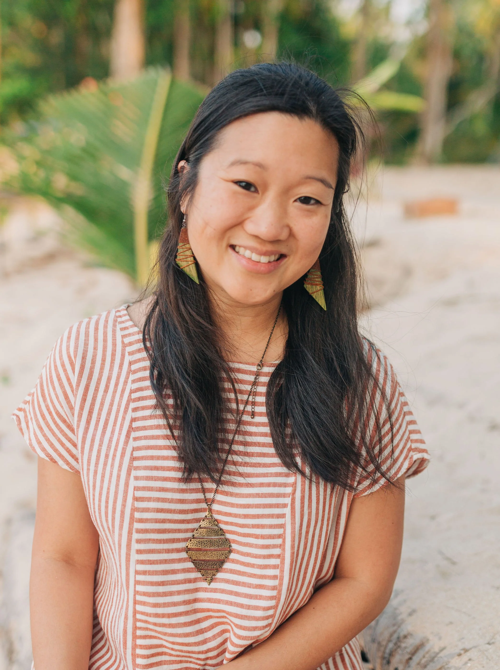 A smiling woman with dark hair wearing a striped shirt, large green leaf earrings, and a pendant necklace, outdoors with blurred greenery in the background.