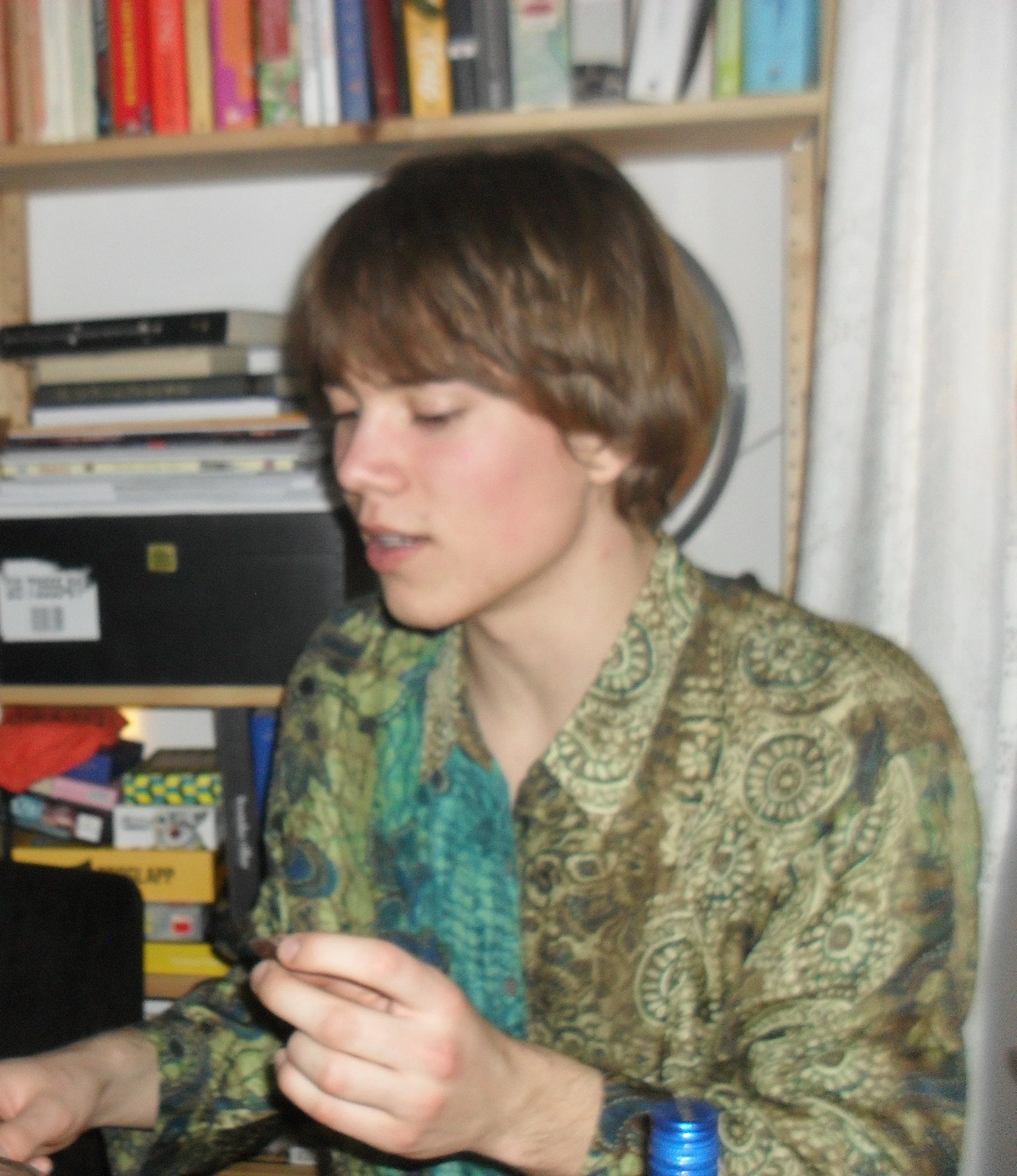 A person with short brown hair wearing a green paisley shirt, looking down at a smartphone in their hand. In the background, there is a bookshelf filled with books and shelves to the right.