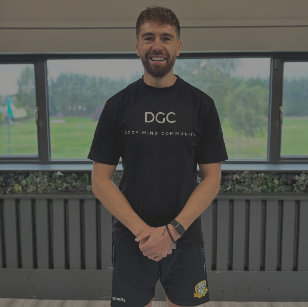 A smiling man with a beard and short curly hair standing indoors in front of large windows with a park or field view. He is wearing a black T-shirt that reads 'DGC Body Mind Community' and black shorts with a patch on the left leg.