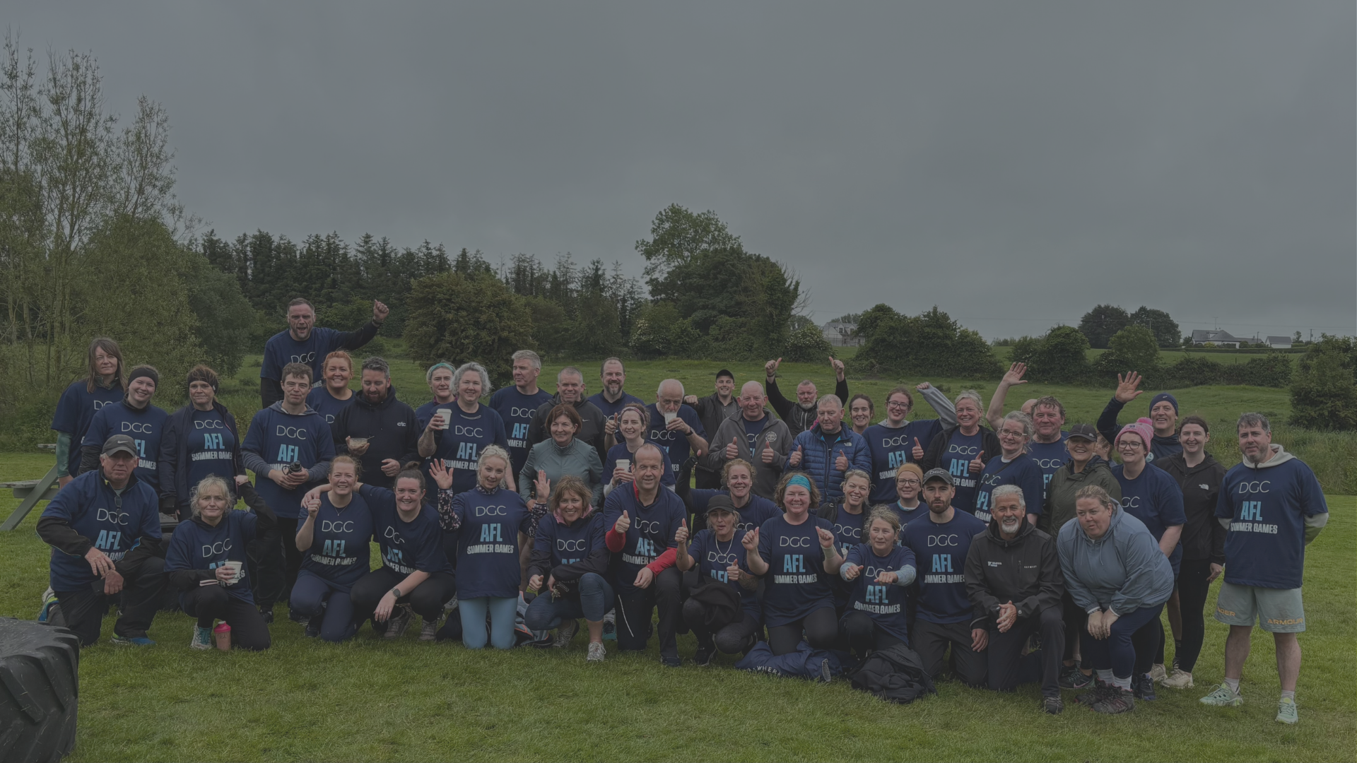 Group of people outdoors wearing matching navy blue T-shirts with AFL Summer Games logo, smiling and posing for a photo on a grassy field with trees and overcast sky in the background.