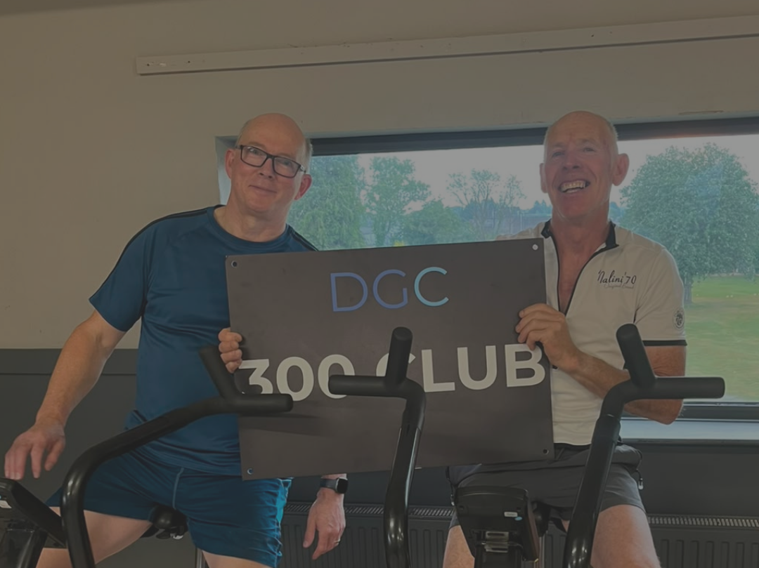 Two older men at a gym holding a sign that reads 'DGC ZOO CLUB'. One man is wearing a blue athletic shirt and glasses, and the other is in a white shirt with a logo and shorts, both smiling. Exercise equipment is visible.