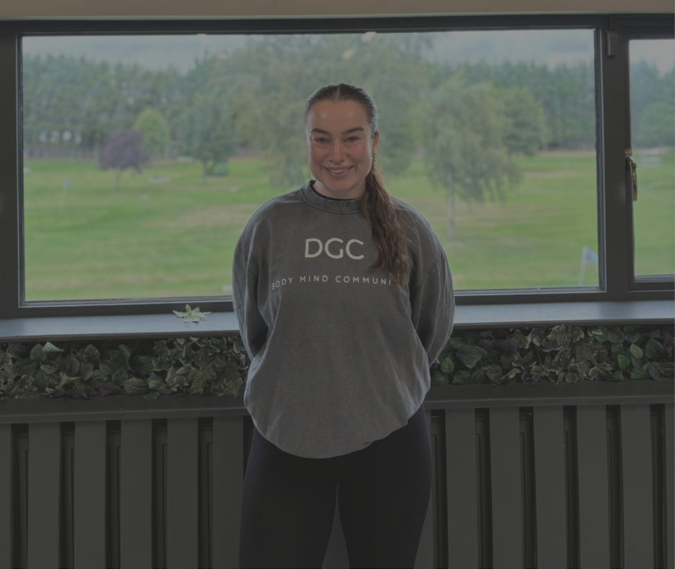 A young woman with a ponytail smiling inside a room with large windows overlooking a green park.