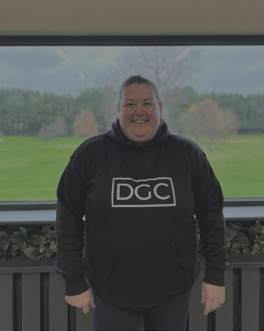 A smiling woman standing indoors in front of a large window with a view of trees and grass outside, wearing a black hoodie with 'DGC' written on it.