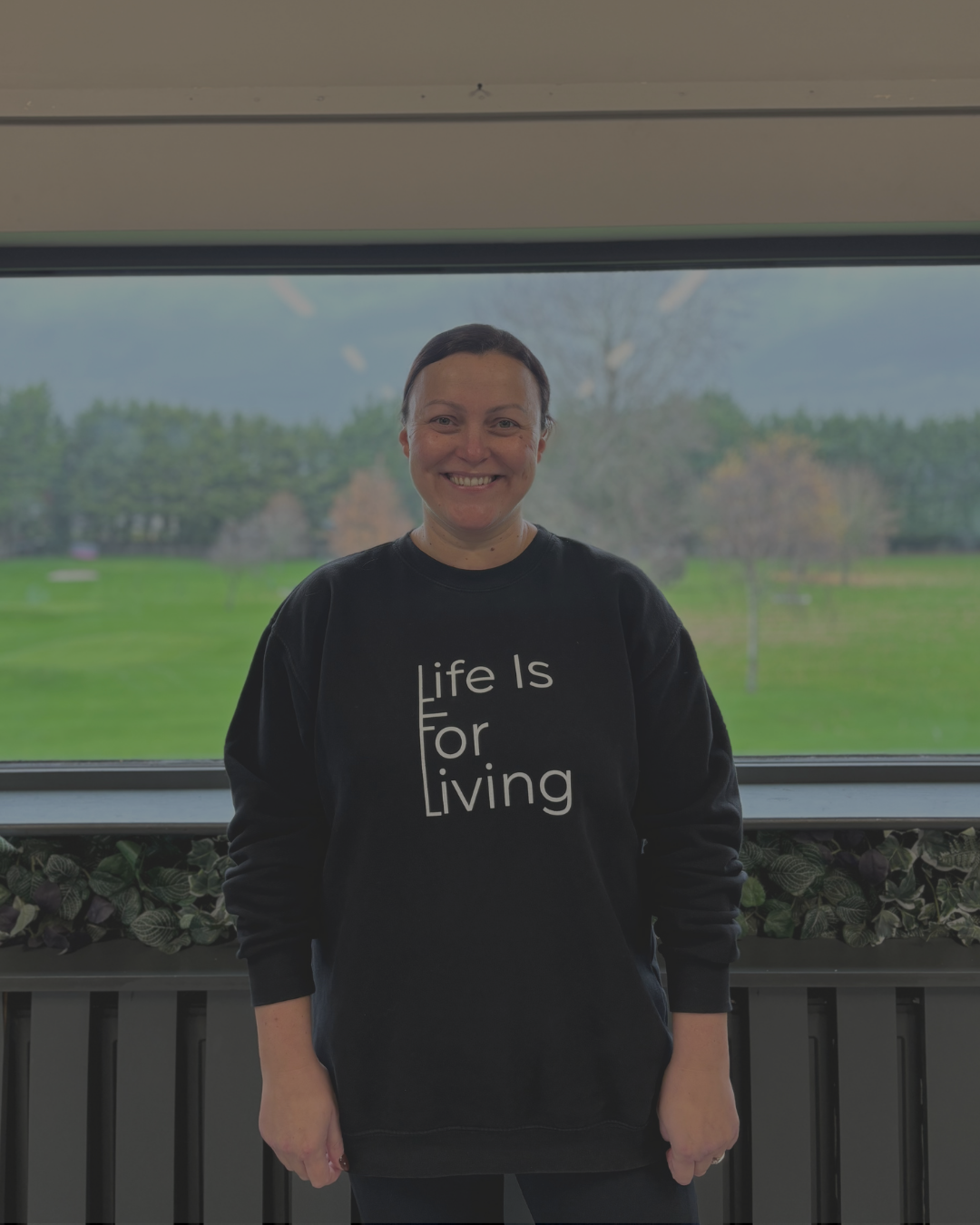 A smiling person standing indoors in front of a large window with a view of a green park and trees outside. They are wearing a black sweatshirt that says "Life Is For Living" in white letters.