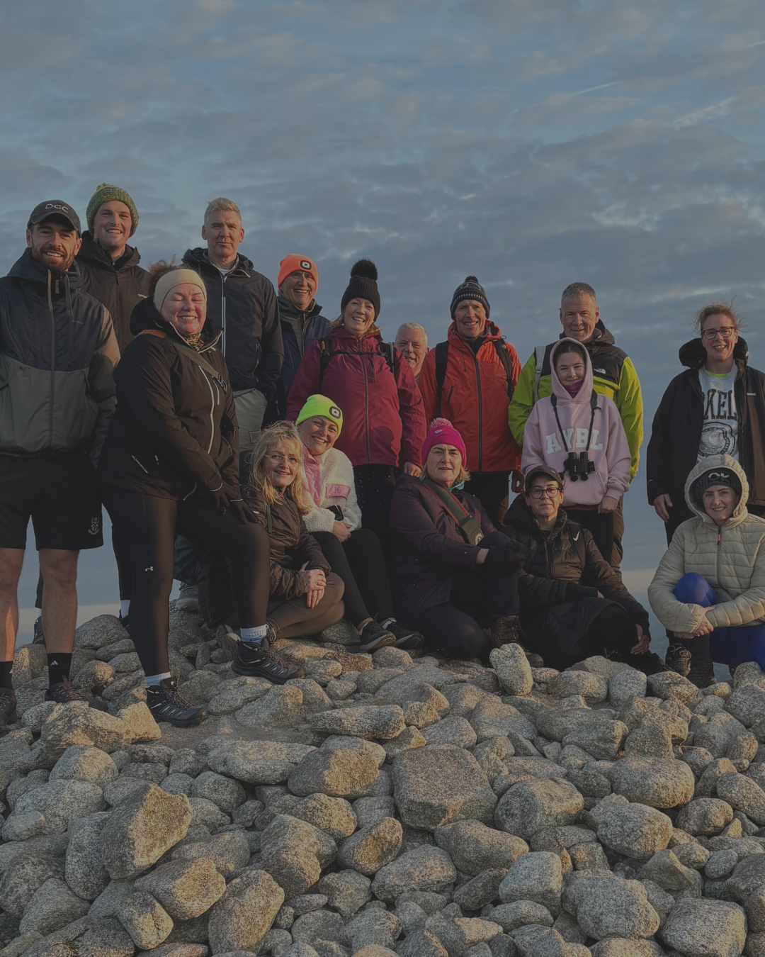Group of people, dressed in outdoor and winter clothing, gathered on a rocky hilltop at sunset or sunrise.