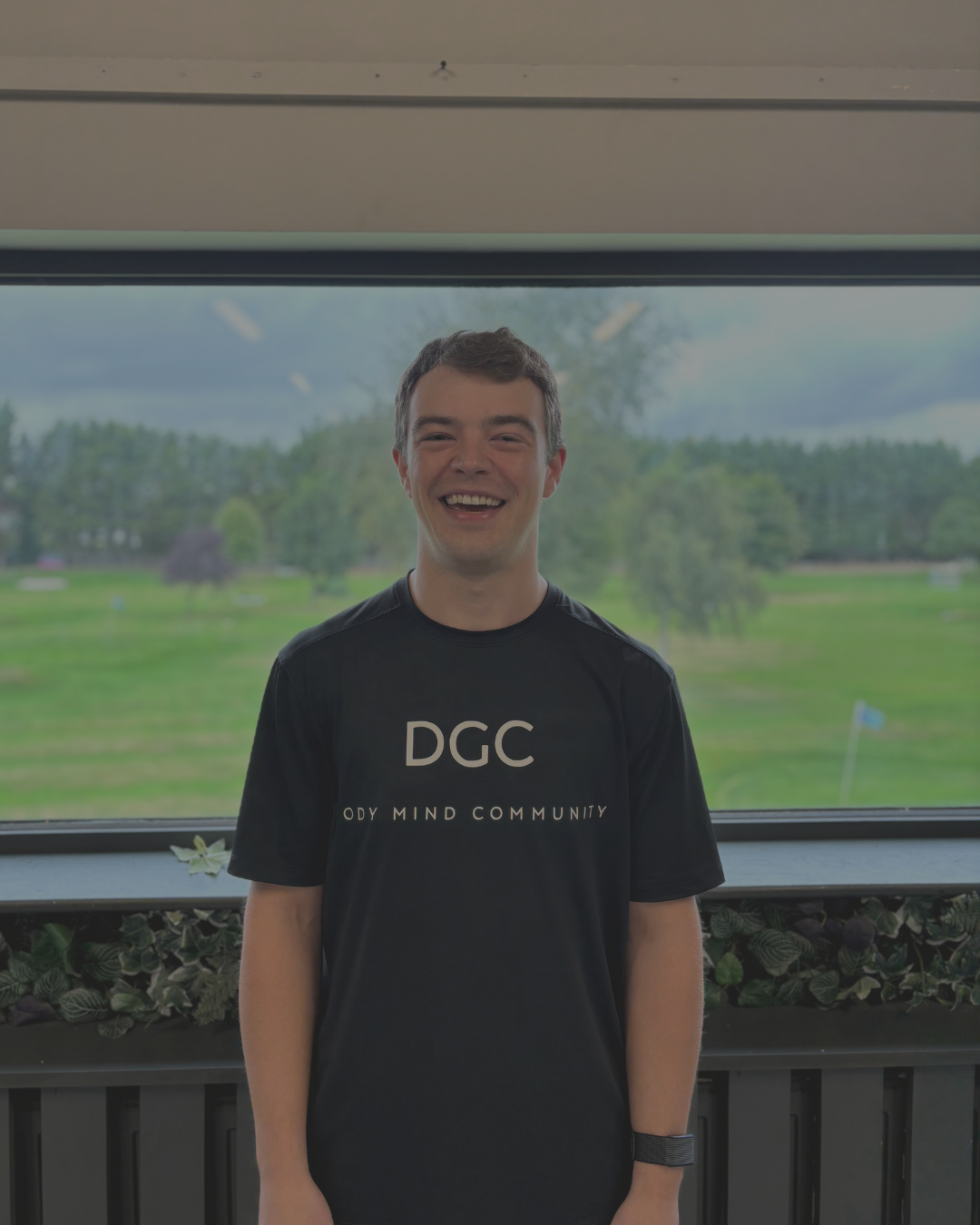 Young man smiling, standing indoors in front of a large window with a view of a golf course and trees outside.