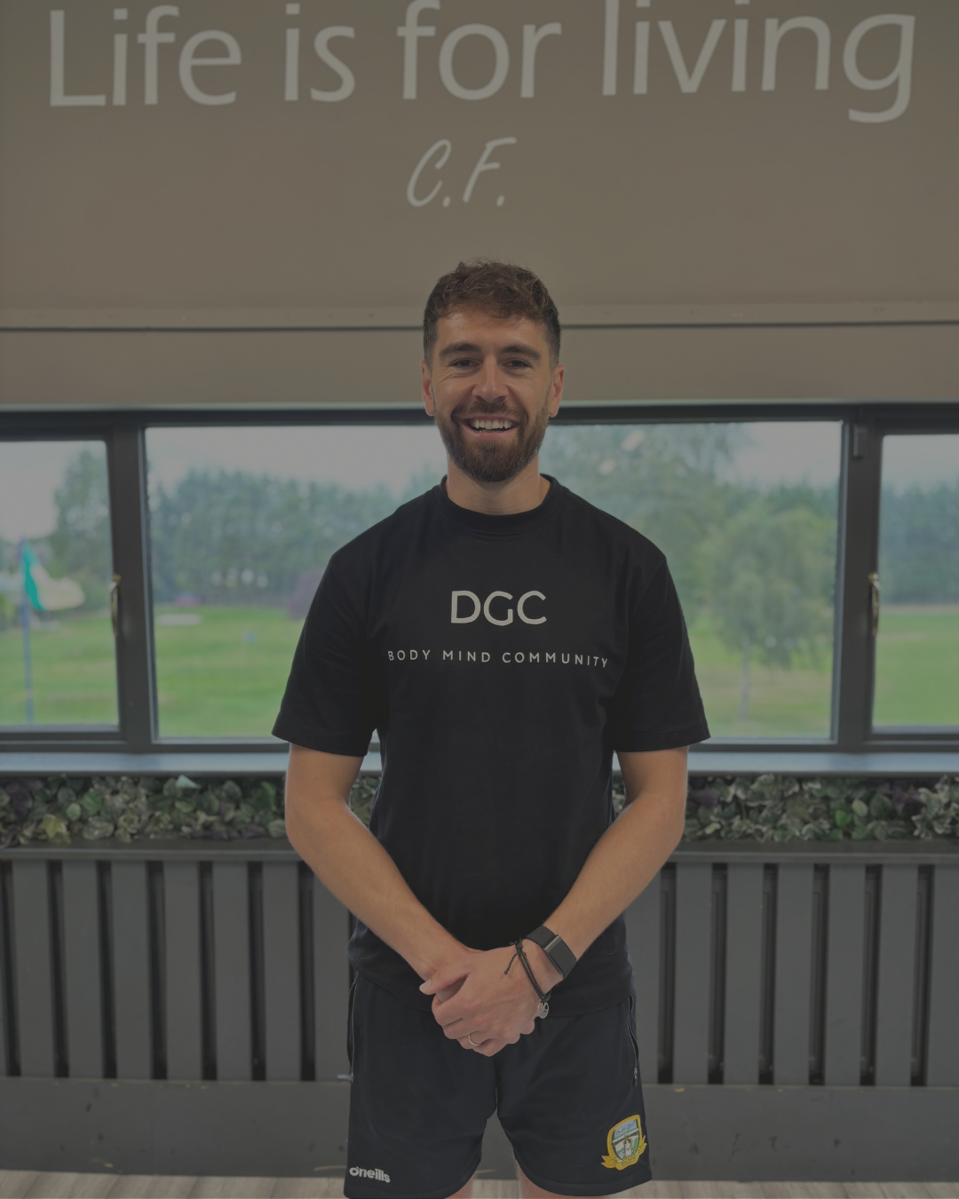 A young man with a beard smiling, standing indoors with windows showing a green outdoor landscape, wearing a black T-shirt with 'DGC Body Mind Community' and black shorts.