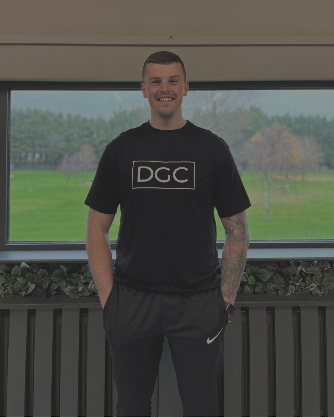 A young man with short brown hair and tattoos on his left arm, wearing a black t-shirt with 'DGC' printed on it and black Nike pants, standing indoors in front of a large window overlooking a grassy park with trees.