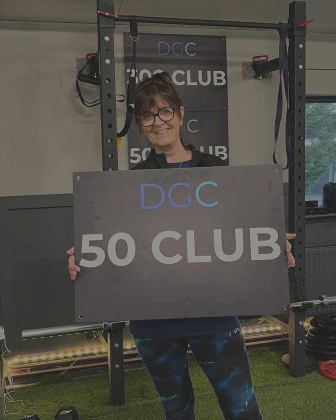 A smiling woman wearing glasses holding a large sign that reads 'DGC 50 CLUB' in a gym with workout equipment in the background.