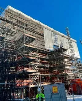 Byggarbetsplats med scaffolding och byggnadsdela, en byggnad under konstruktion, mot en klar blå himmel.