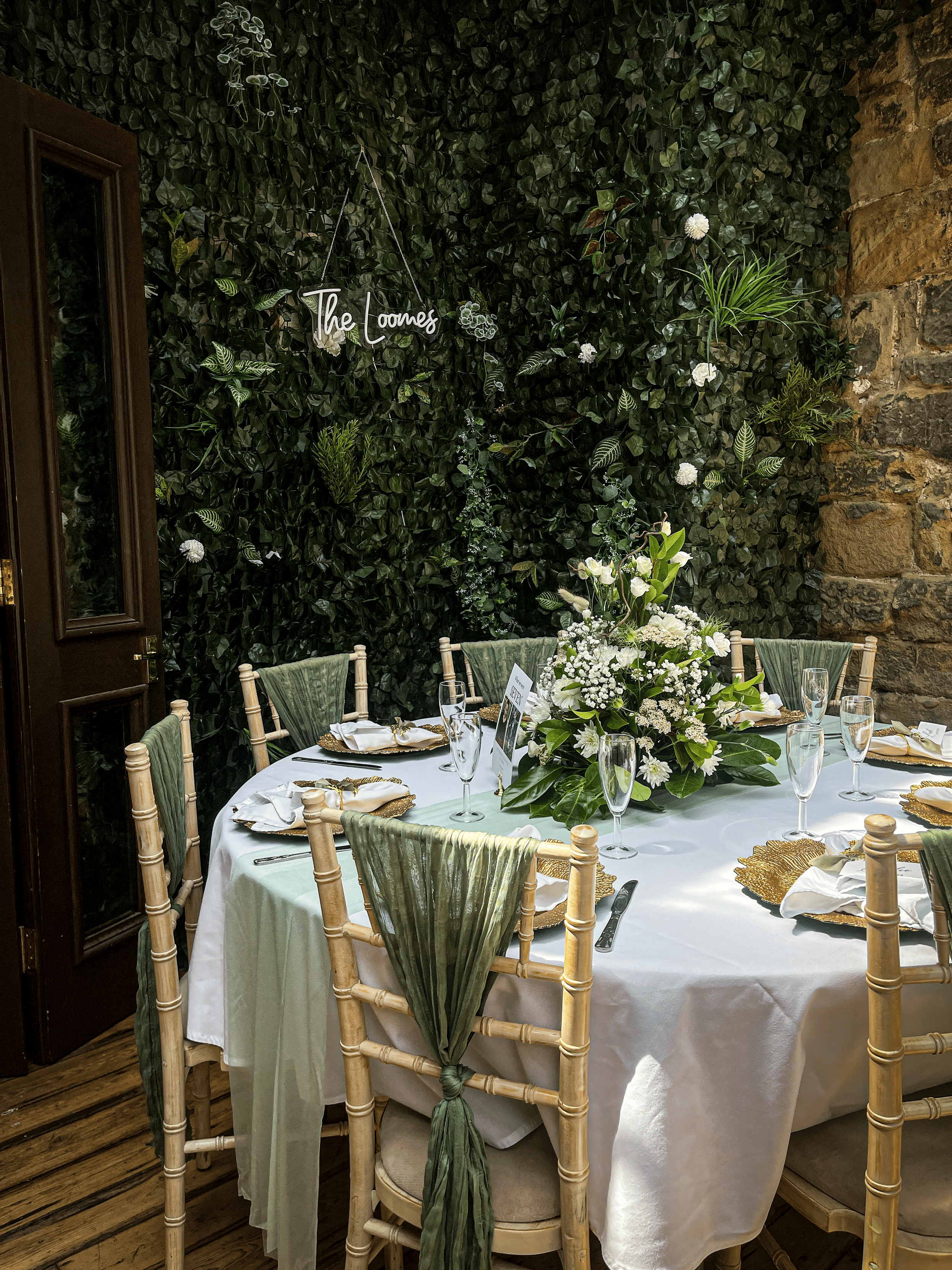 A round dining table set for a formal event with white tablecloth, gold chargers, white napkins, and glasses, decorated with a large floral arrangement of white flowers and greenery. The setting is in a room with a green leafy wall, a stone wall on the right, and wooden flooring. There is a sign on the wall that reads "The Loones."