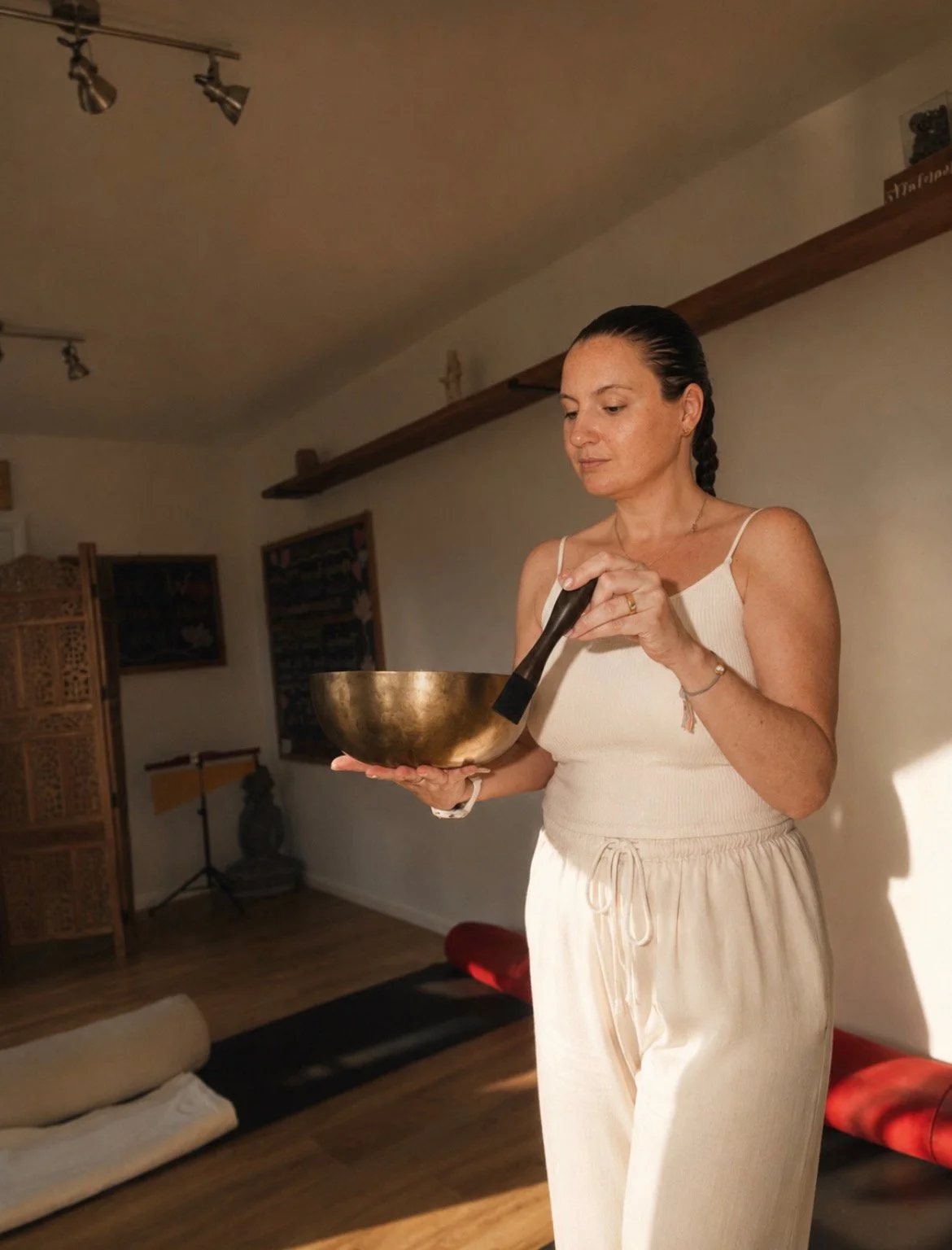 A woman with dark hair in a braid, wearing a white tank top and beige sweatpants, stands indoors holding a large bell-shaped singing bowl and a wooden mallet, with a peaceful expression.