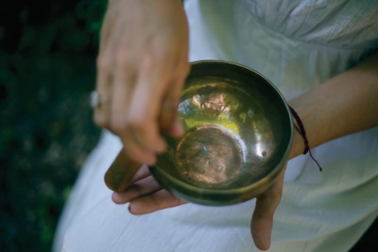 Person holding a singing bowl with a wooden striker, outdoors with greenery in the background.