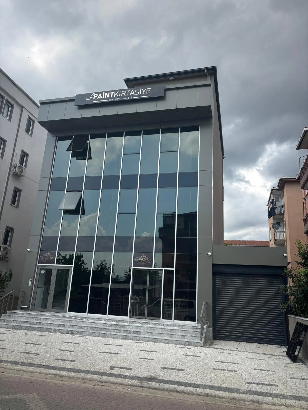 Modern glass-fronted building with dark exterior, steps at the entrance, and cloudy sky