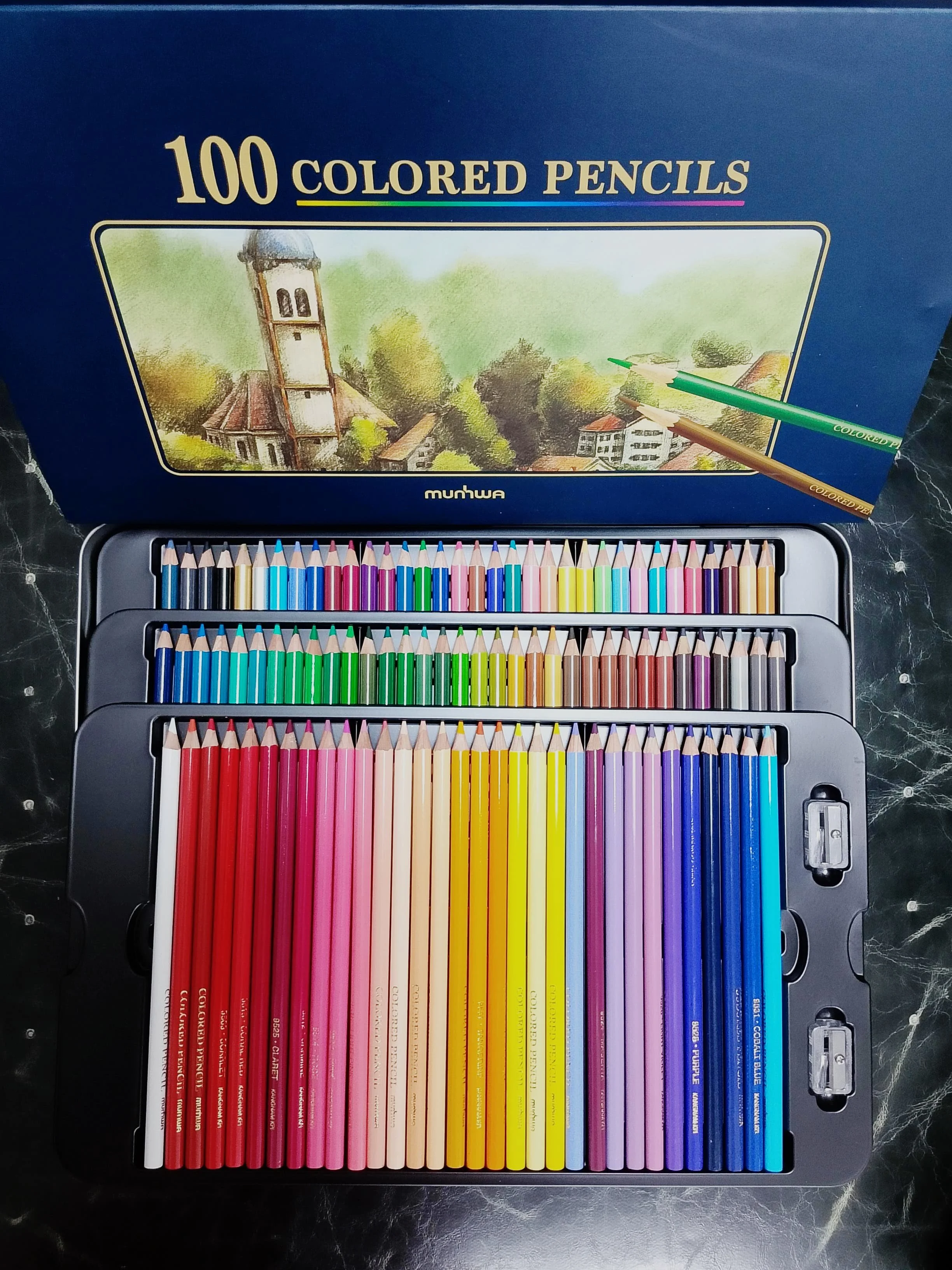 A blue box of 100 colored pencils with an illustration of a village scene on the lid, and a tray of rainbow-colored pencils arranged in a gradient order below.