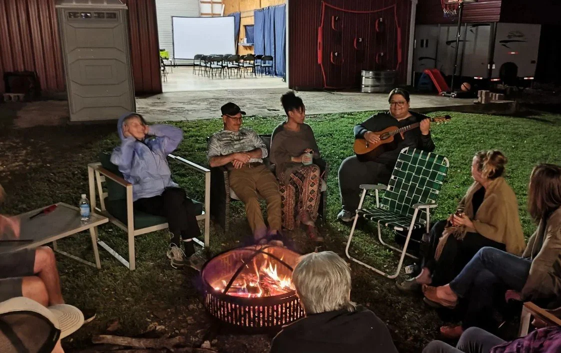 People sitting around a campfire at night, with a person playing guitar, in an outdoor area with a barn or warehouse in the background.