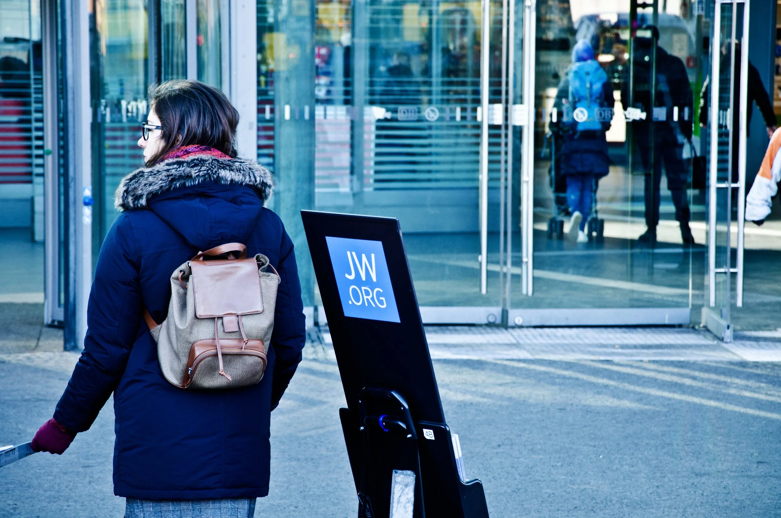 Woman with glasses and a backpack waiting outside an airport gate, holding a rolling suitcase, in a cold-weather jacket with a fur-lined hood, in front of glass doors and a sign for JW.org.