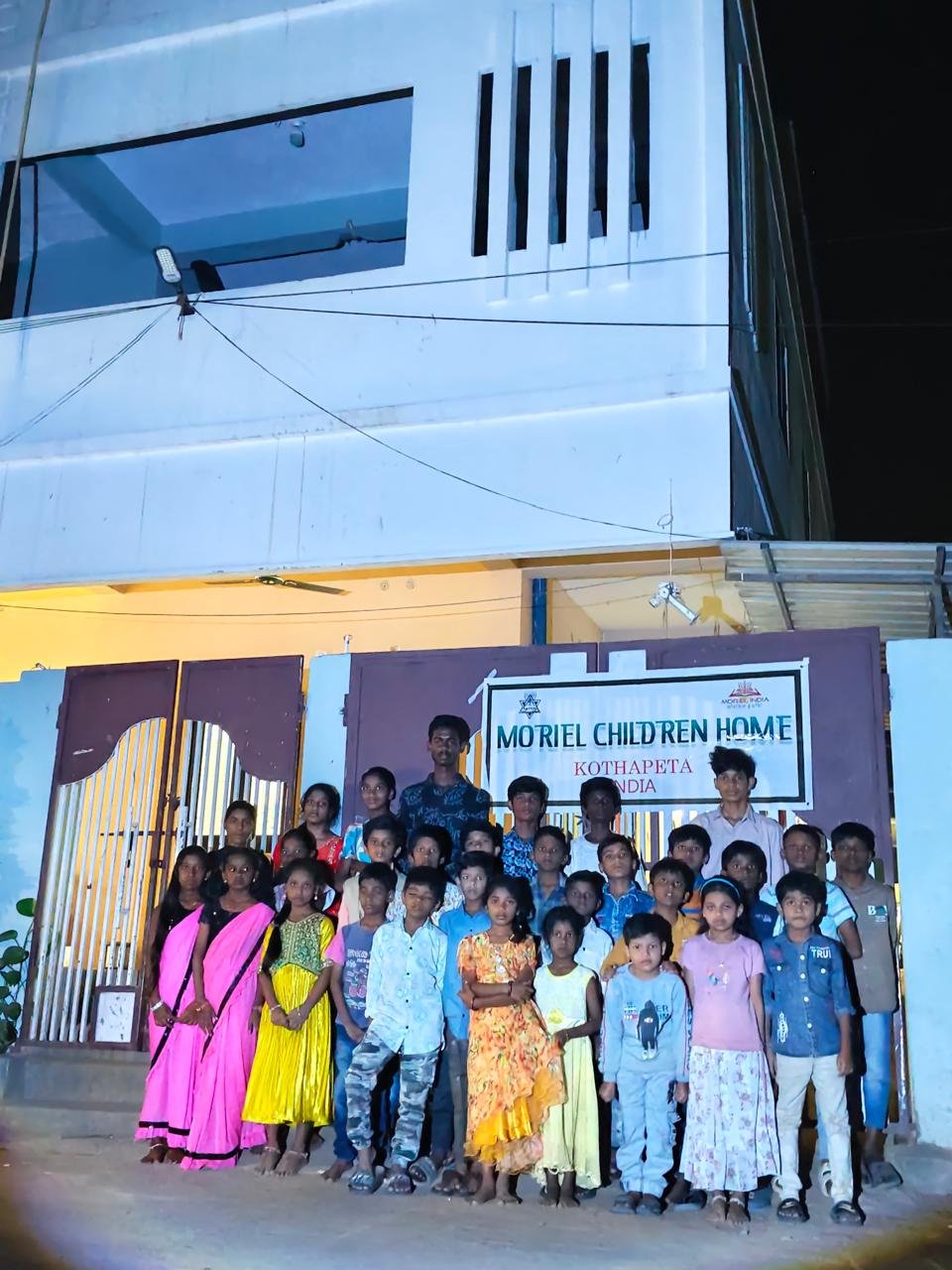 Children and adults standing in front of a sign that reads 'MORIEL CHILDREN HOME' at night.