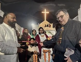 A man shaking hands with another man at a religious event, with a church and cross illuminated in the background, and several women standing nearby.