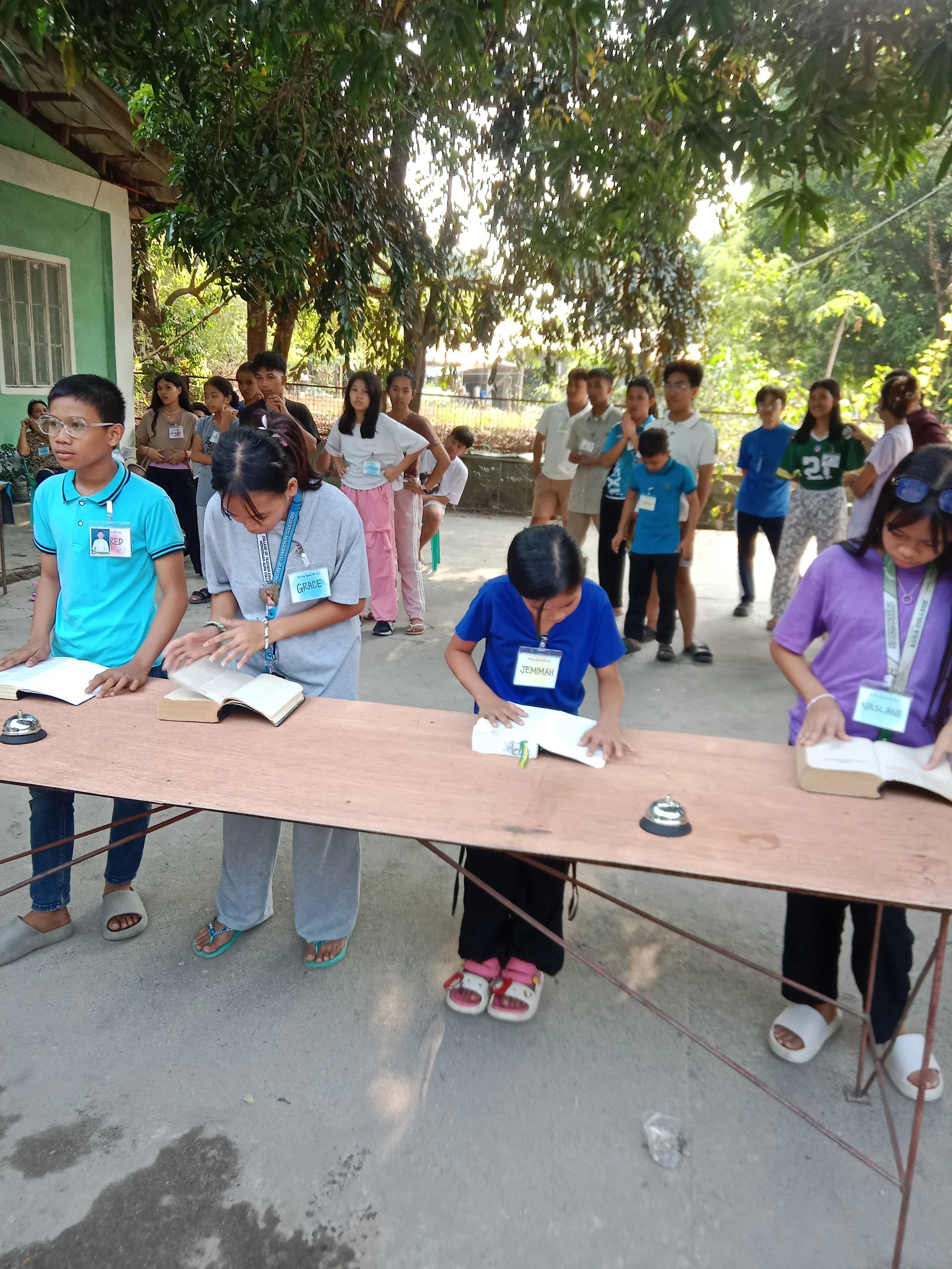 A group of young children and some adults outdoors, participating in a reading activity with books on a table. The children are standing and looking at the books, some are wearing name tags. The setting seems to be a garden or yard with trees and a small building in the background.