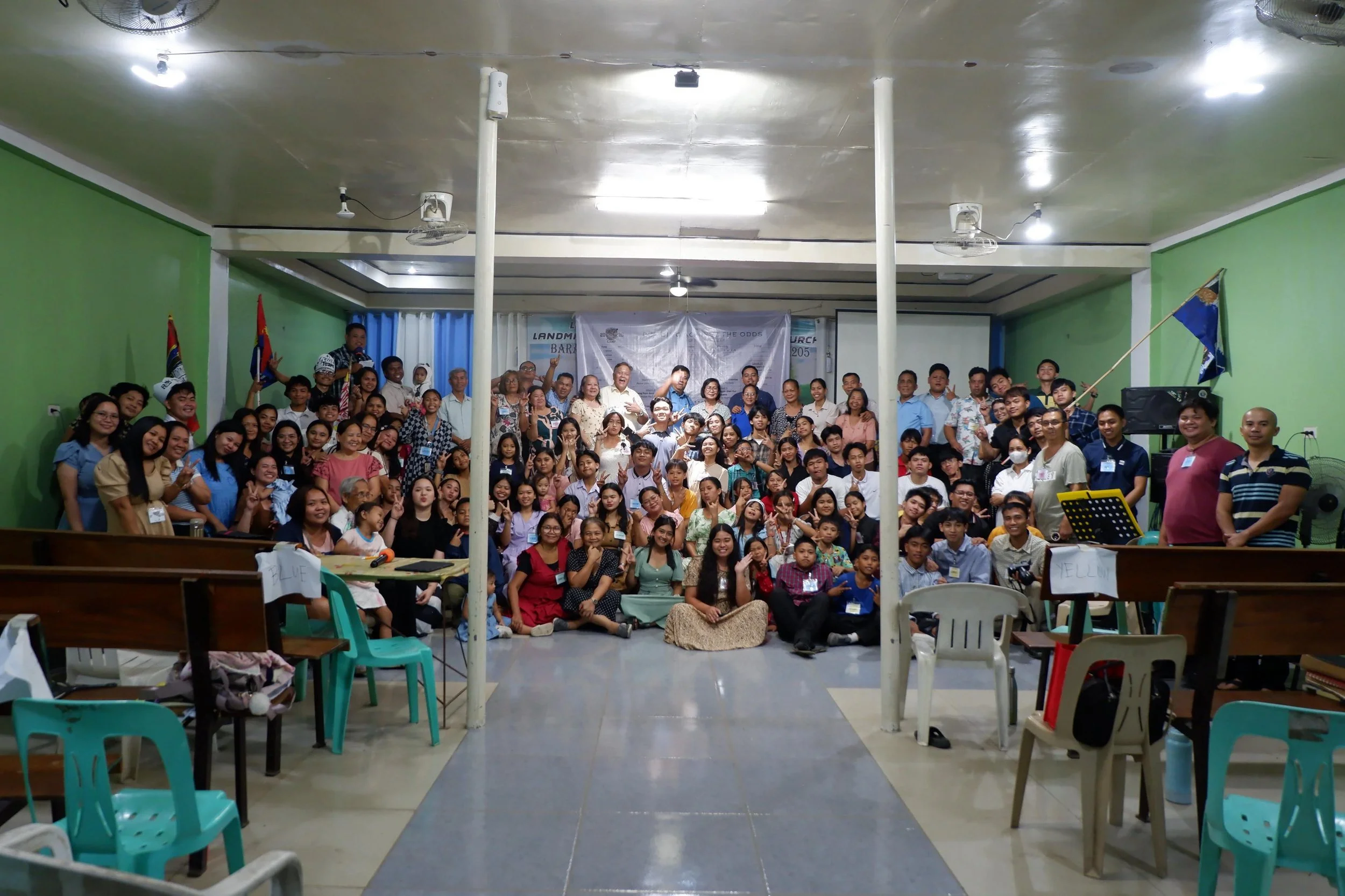 A large group of diverse people gathered in a brightly lit room, posing for a group photo. The room has green walls, flags, and a banner in the background. Some people are smiling and waving, and there are chairs and tables around.