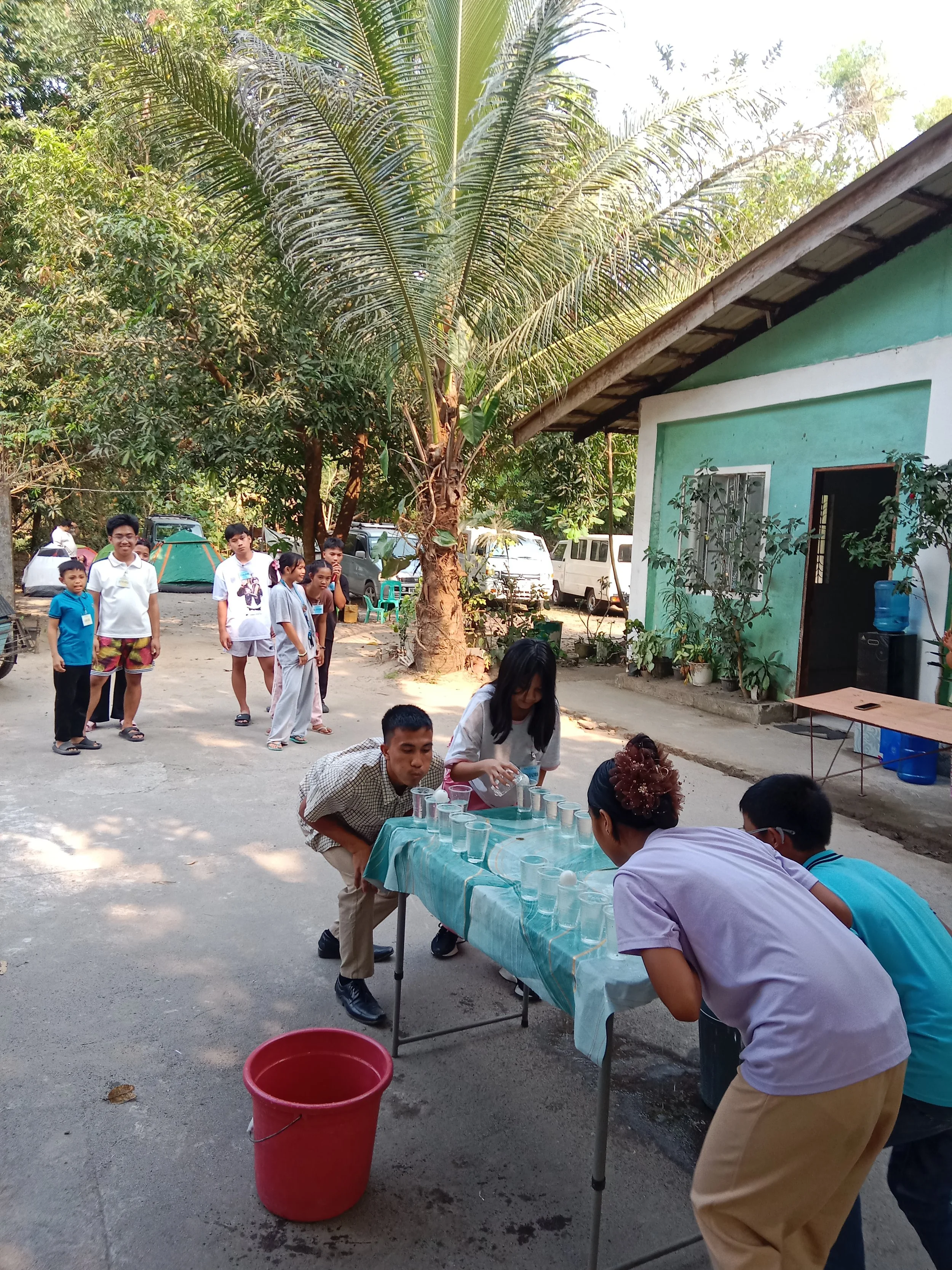 Group of children and adults playing a game outdoors, with glasses and eggs on a table, in a yard with trees and a building visible.