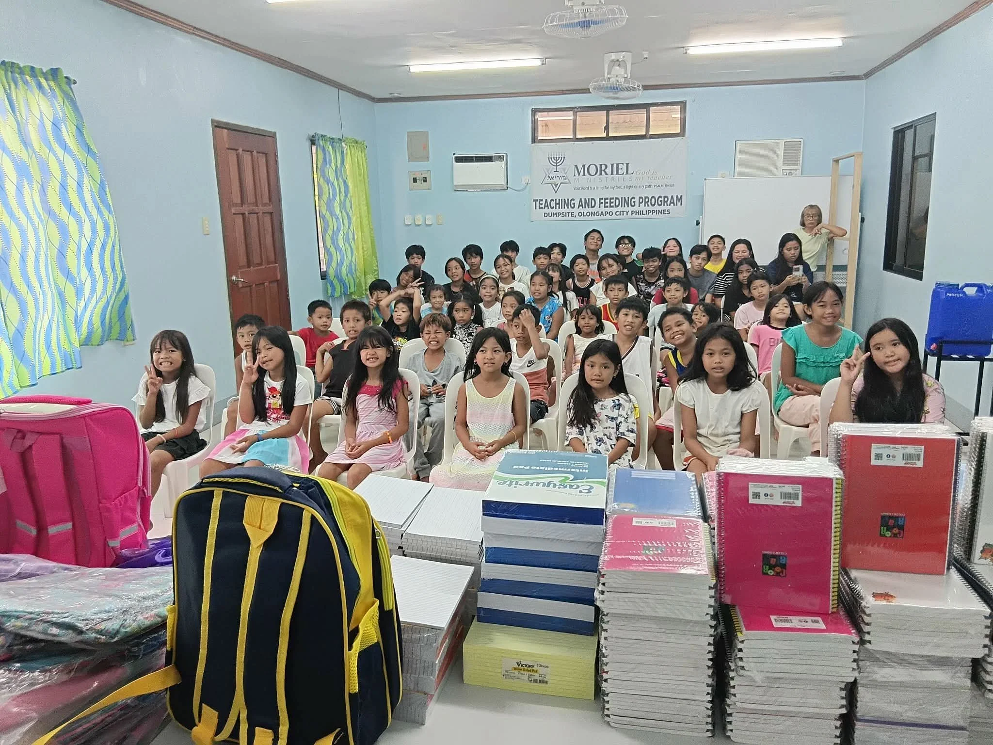A room filled with children sitting on chairs and smiling at the camera. In the background, there's a whiteboard, a banner with religious symbols and text, and colorful curtains. The foreground shows a table with school supplies, books, and backpacks.