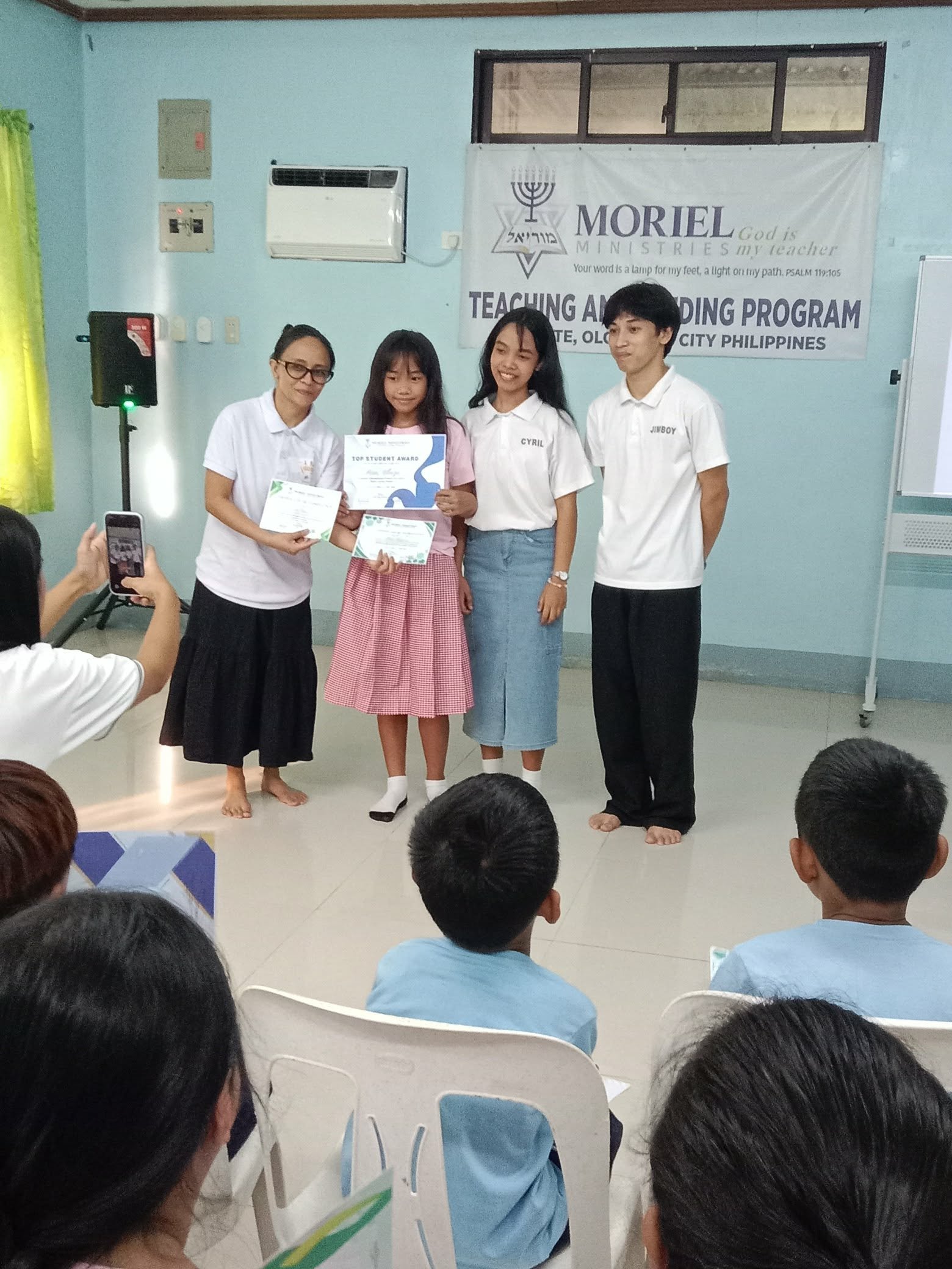 A girl receiving an award in a classroom or community center during a program in Olongapo City, Philippines, with other children and a woman presenting her the award. Some children are seated and watching, with one person taking a photo.