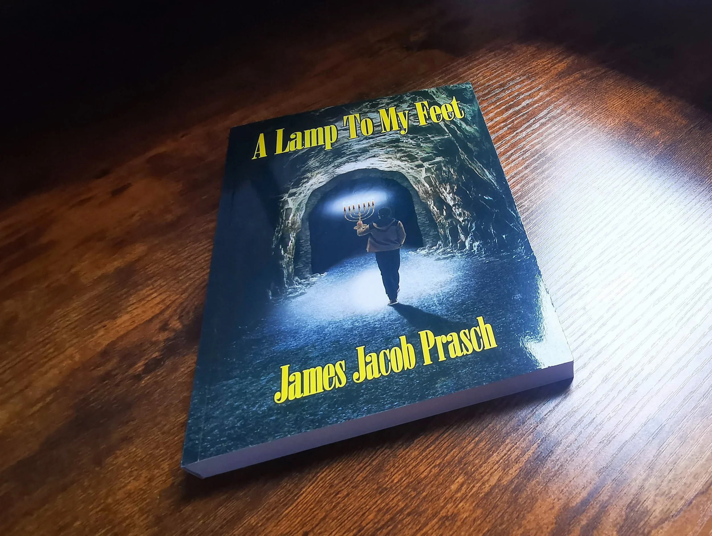 Book cover titled "A Lamp To My Feet" by James Jacob Prasch. The cover features a person holding a menorah inside a dark, tunnel-like cave.