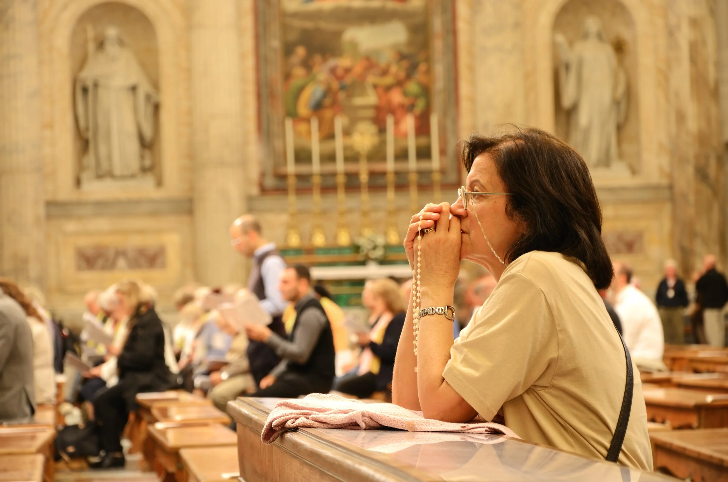 A woman praying with rosary beads in a church during a religious service, with other parishioners and religious artwork in the background.