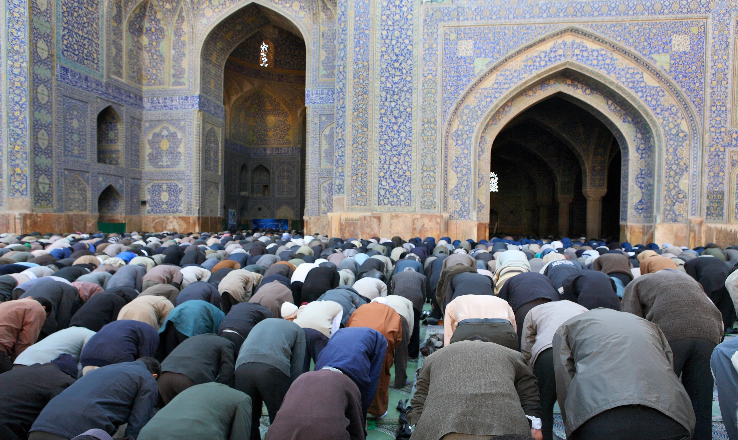 Large group of Muslim men praying inside a decorated mosque, with intricate blue and gold tile work on the walls and arched doorways.
