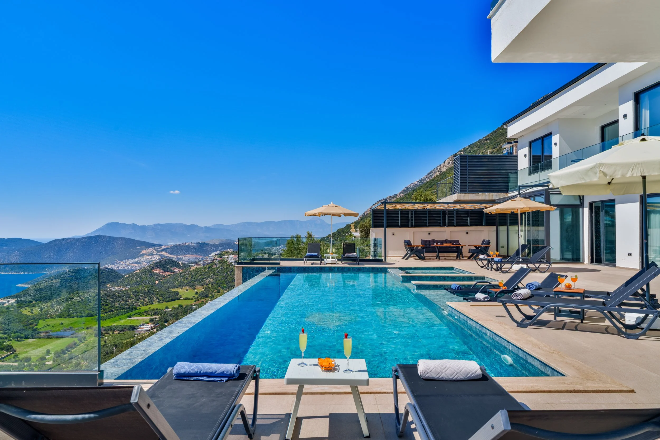 Modern house with balconies, outdoor furniture, umbrellas, and a swimming pool overlooking mountains and a valley.