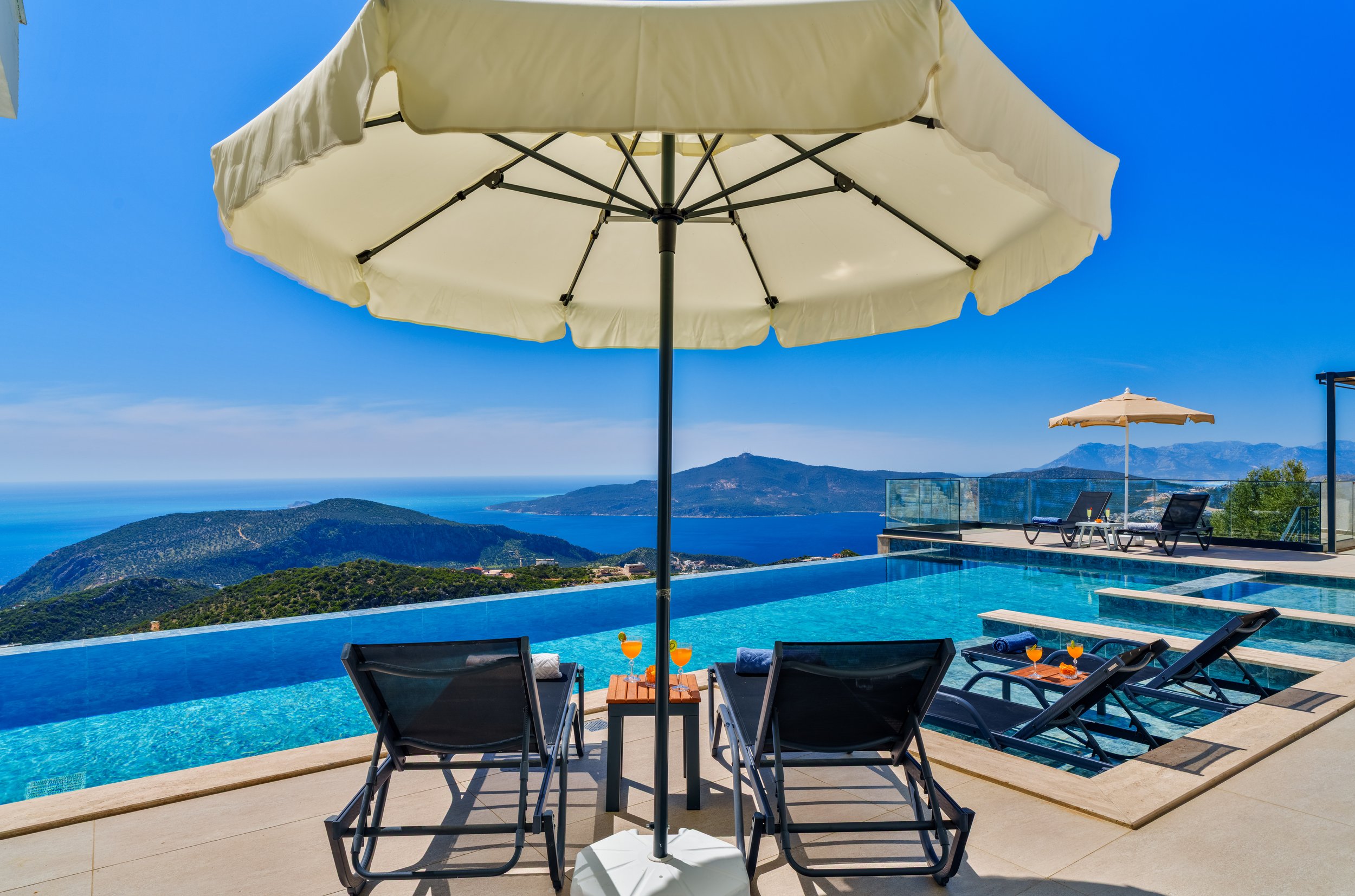 Swimming pool with lounge chairs and umbrellas overlooking mountains and ocean in the distance on a sunny day.