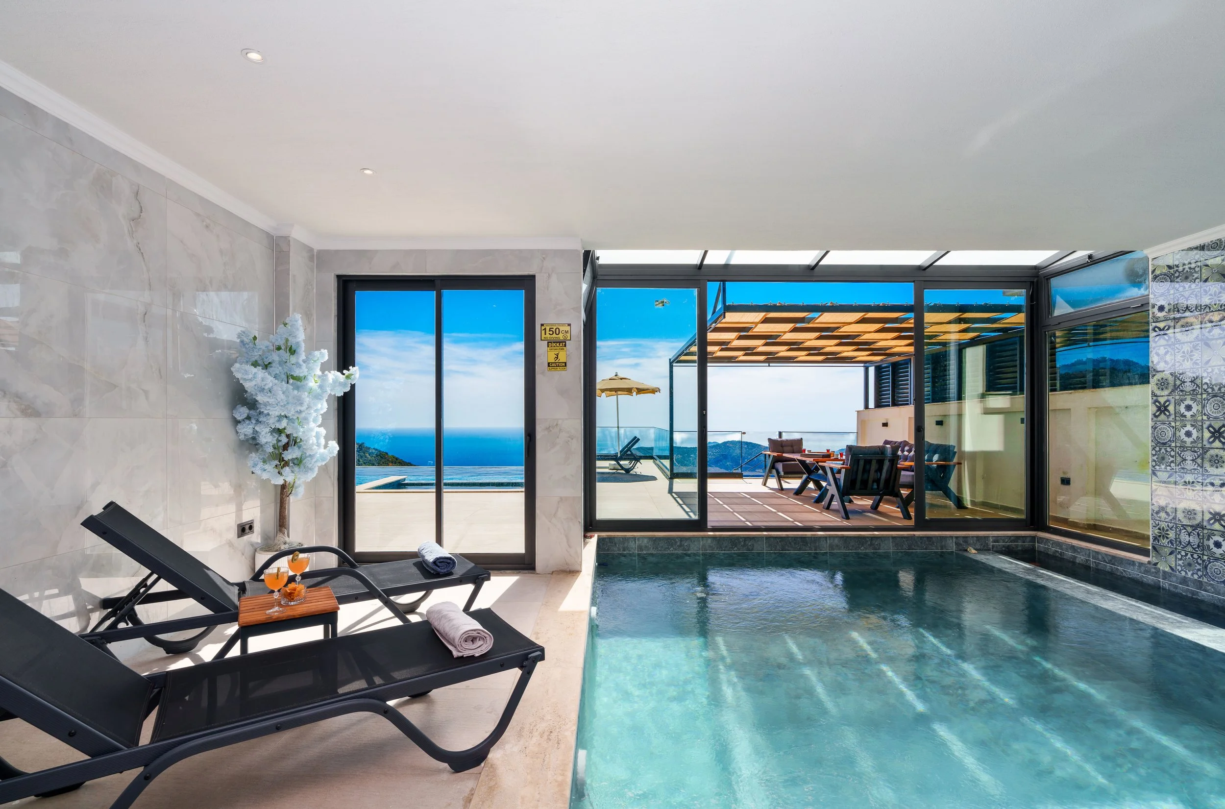 Indoor swimming pool area with lounge chairs, a side table with drinks, a view of an outdoor patio with seating, umbrella, and ocean scenery beyond.