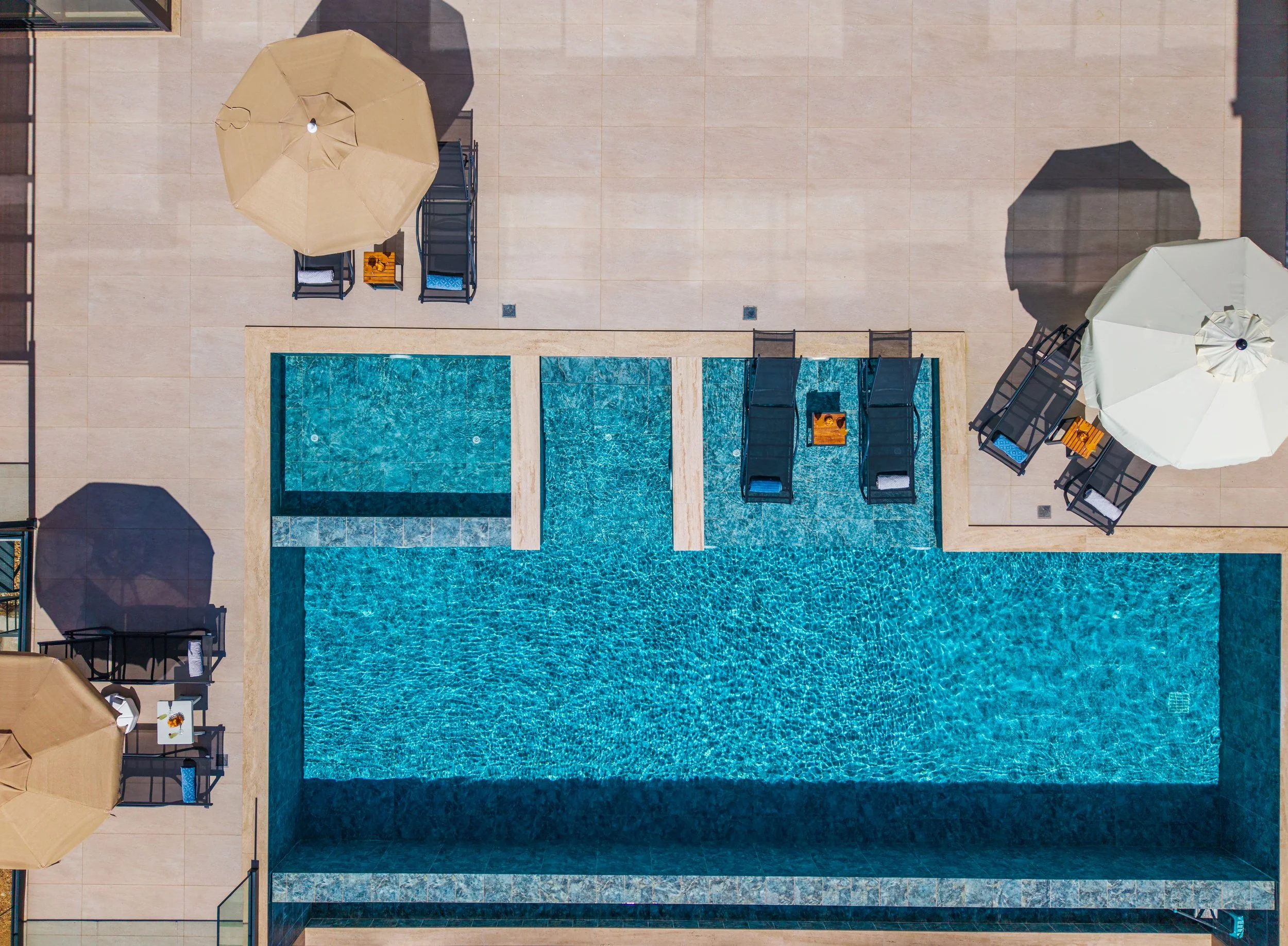 An aerial view of a swimming pool area with beige lounge chairs and umbrellas on a tiled surface.