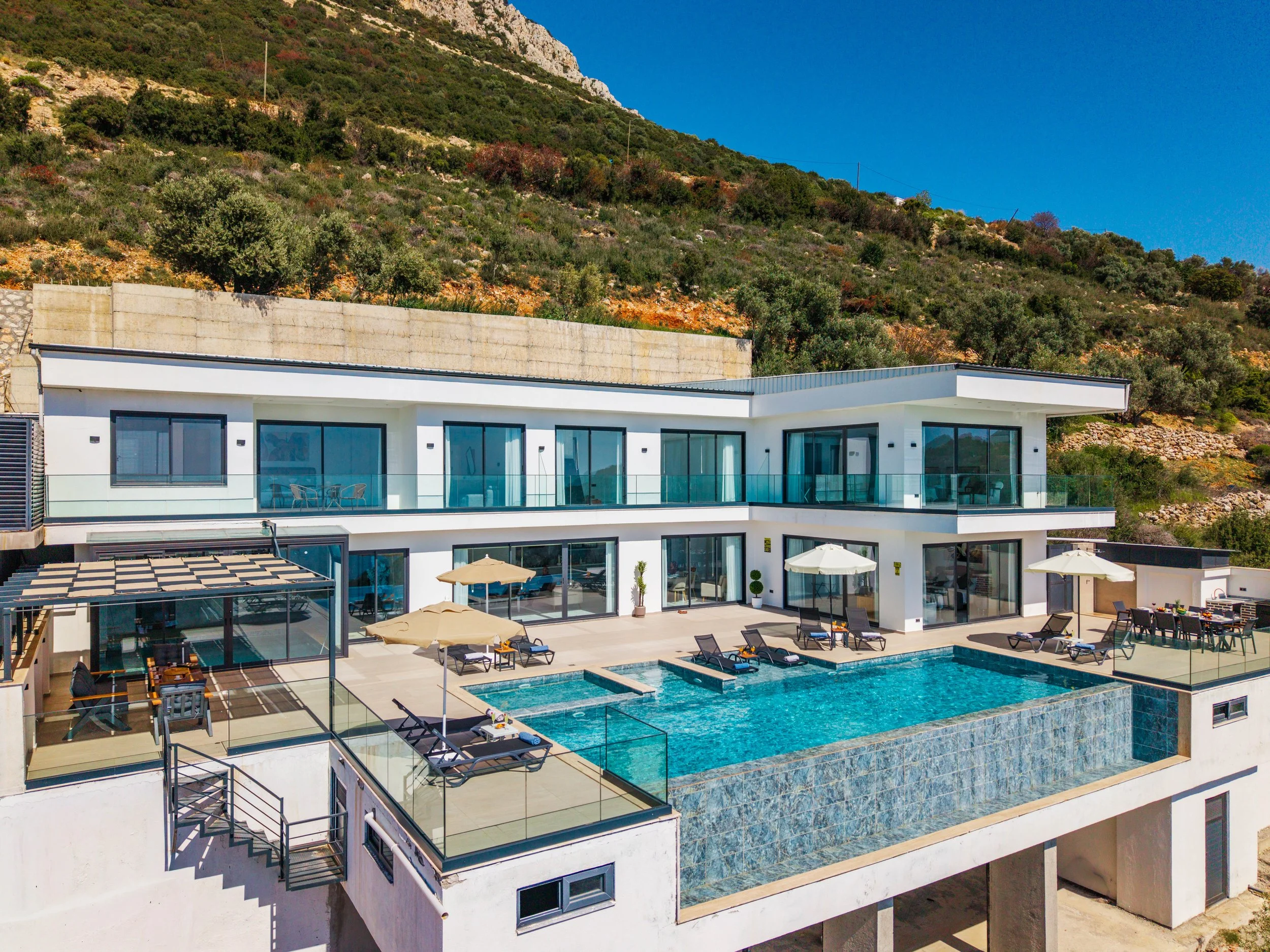 Modern multi-story house with large glass windows and a swimming pool in the backyard, overlooking a hillside with green and red foliage and a clear blue sky.