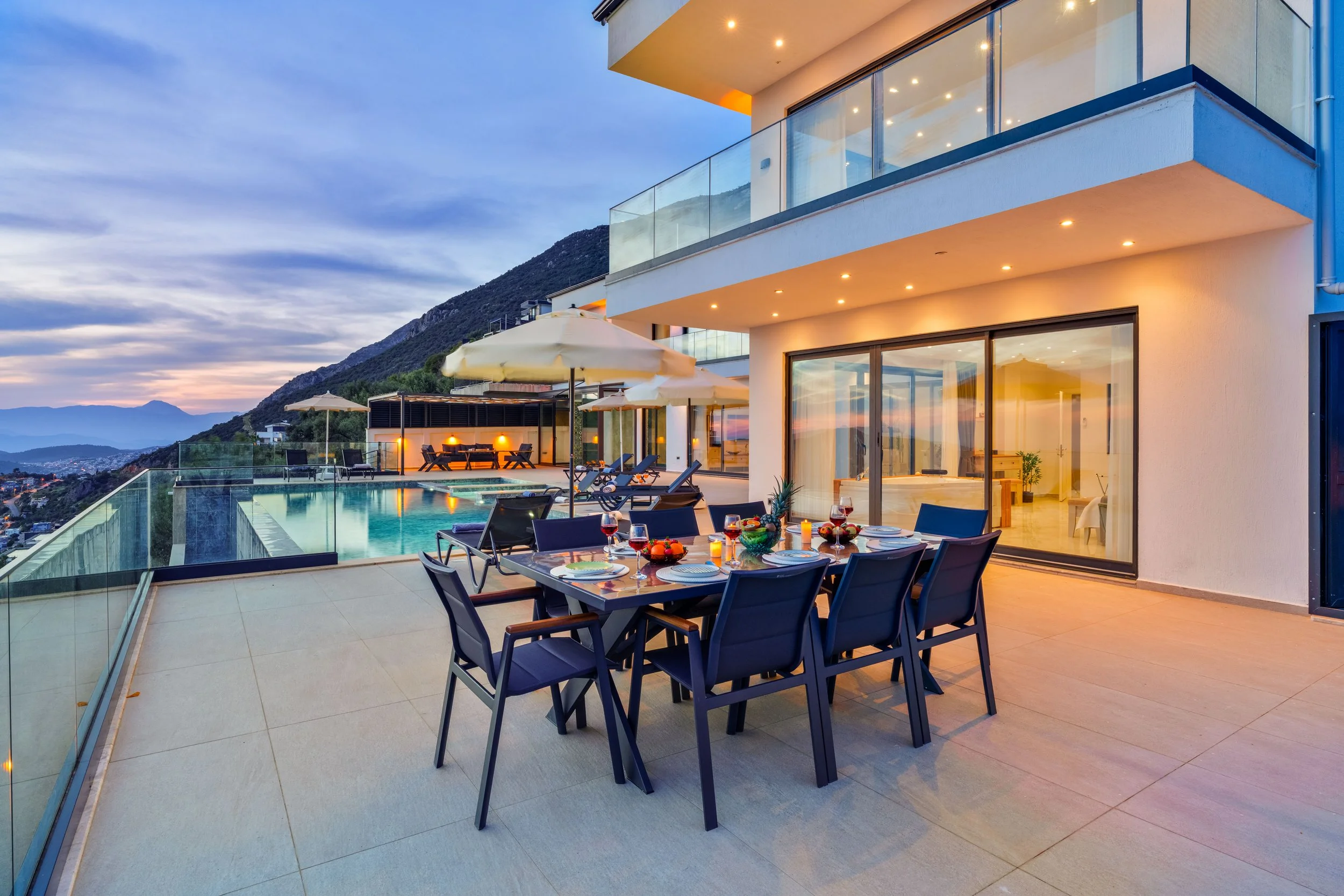 Modern balcony with outdoor dining area overlooking mountains at sunset, pool with glass railing, and lounge chairs with umbrellas.