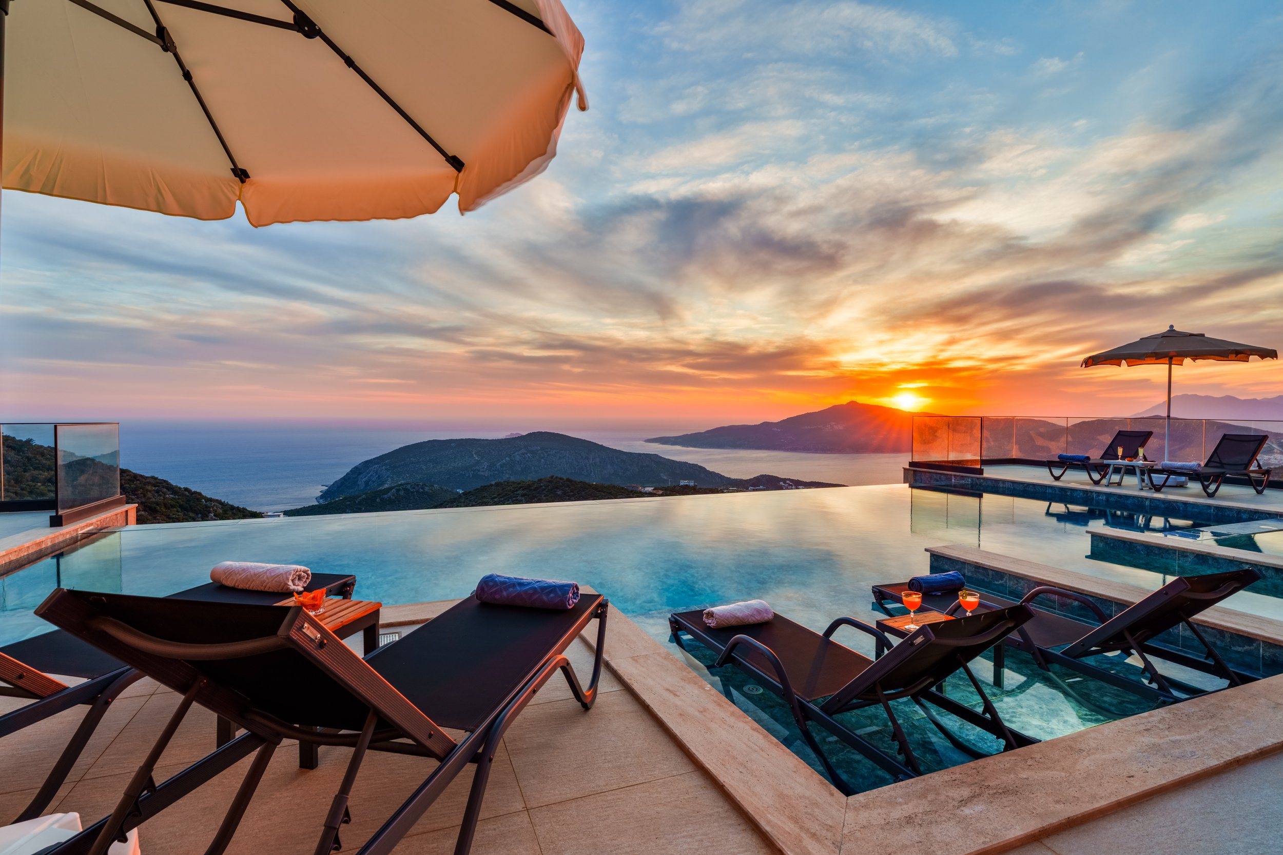 A luxurious infinity pool with a view of mountains and the ocean during sunset, surrounded by lounge chairs and umbrellas.