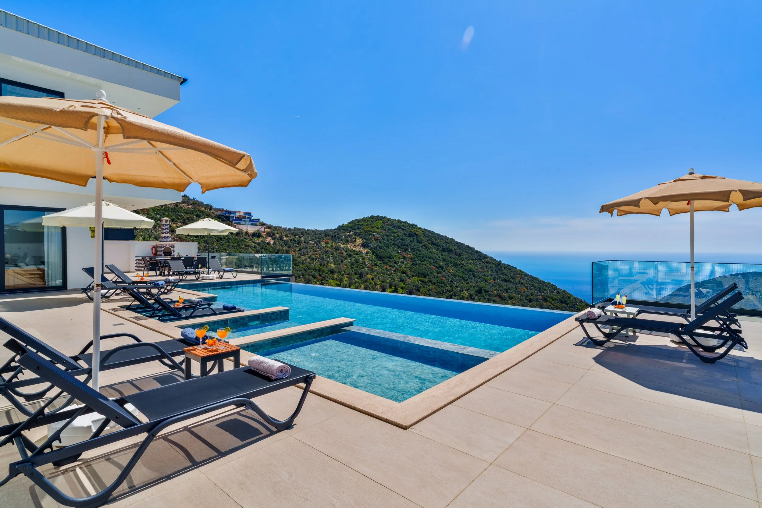 Luxury poolside area with sun loungers, umbrellas, and views of the ocean and hills.