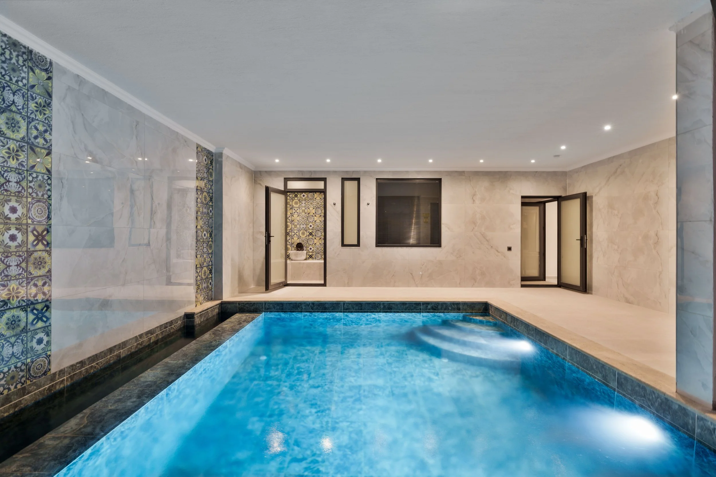 Indoor swimming pool with blue water, surrounded by marble walls and decorated with patterned tiles, with multiple ceiling lights and glass doors and windows.