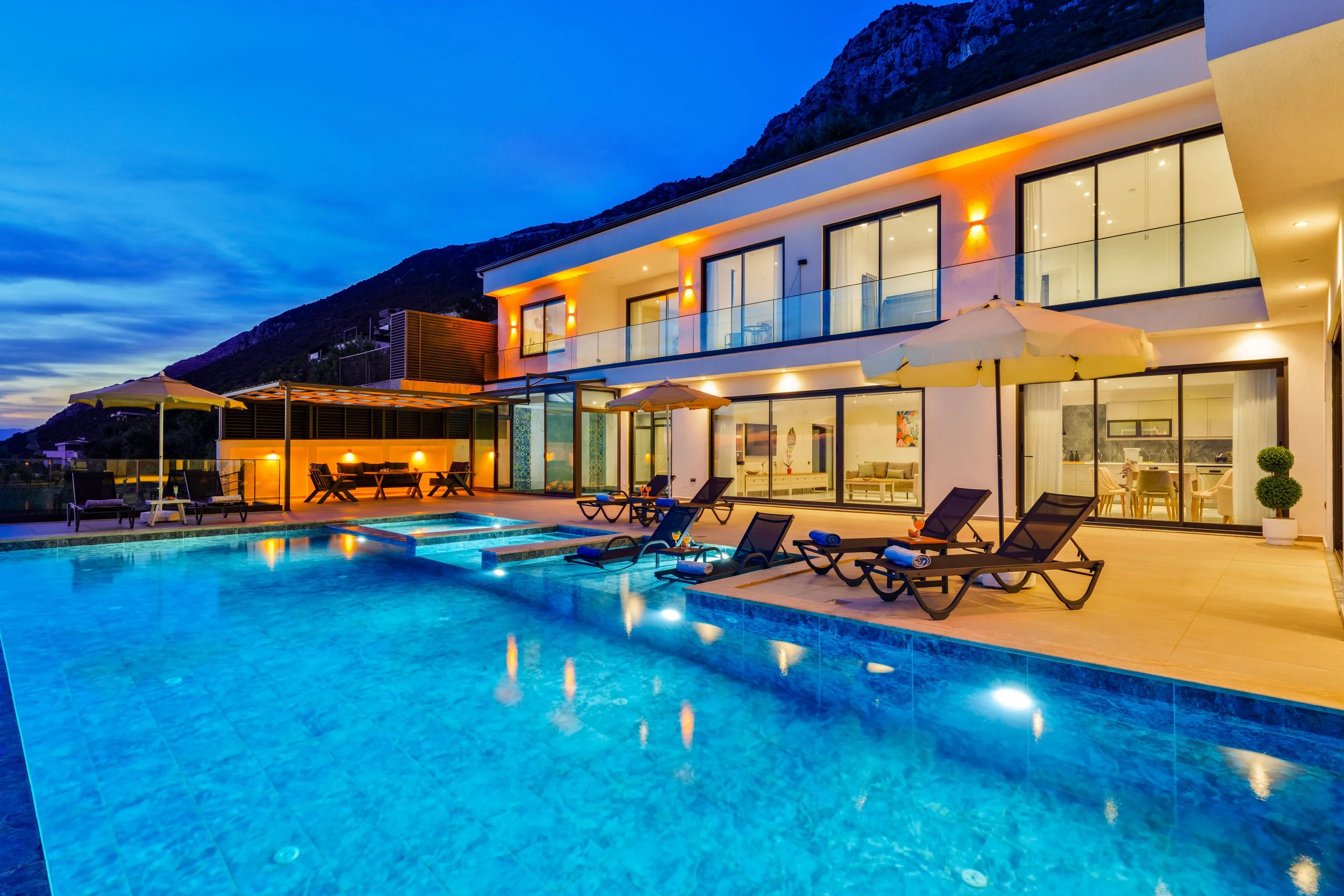 Modern house with large glass windows and balcony, illuminated by warm outdoor lighting, set against a mountainous landscape during dusk, with a swimming pool and lounge chairs on the patio.