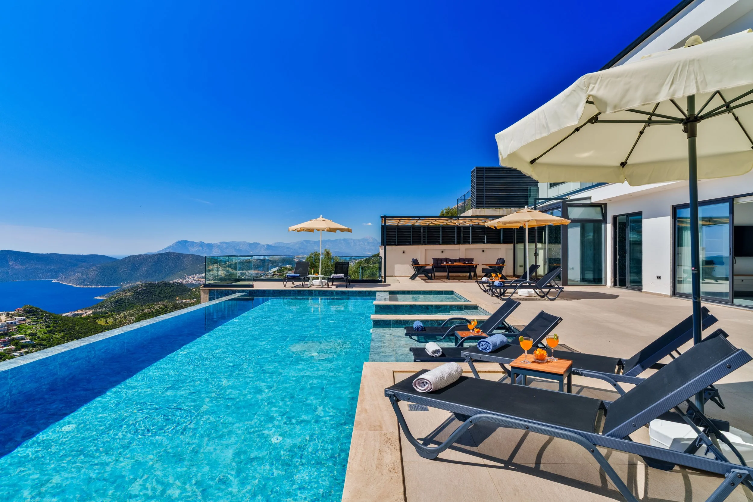 View of a luxury outdoor swimming pool with lounge chairs, umbrellas, and a scenic mountain and ocean landscape in the background.