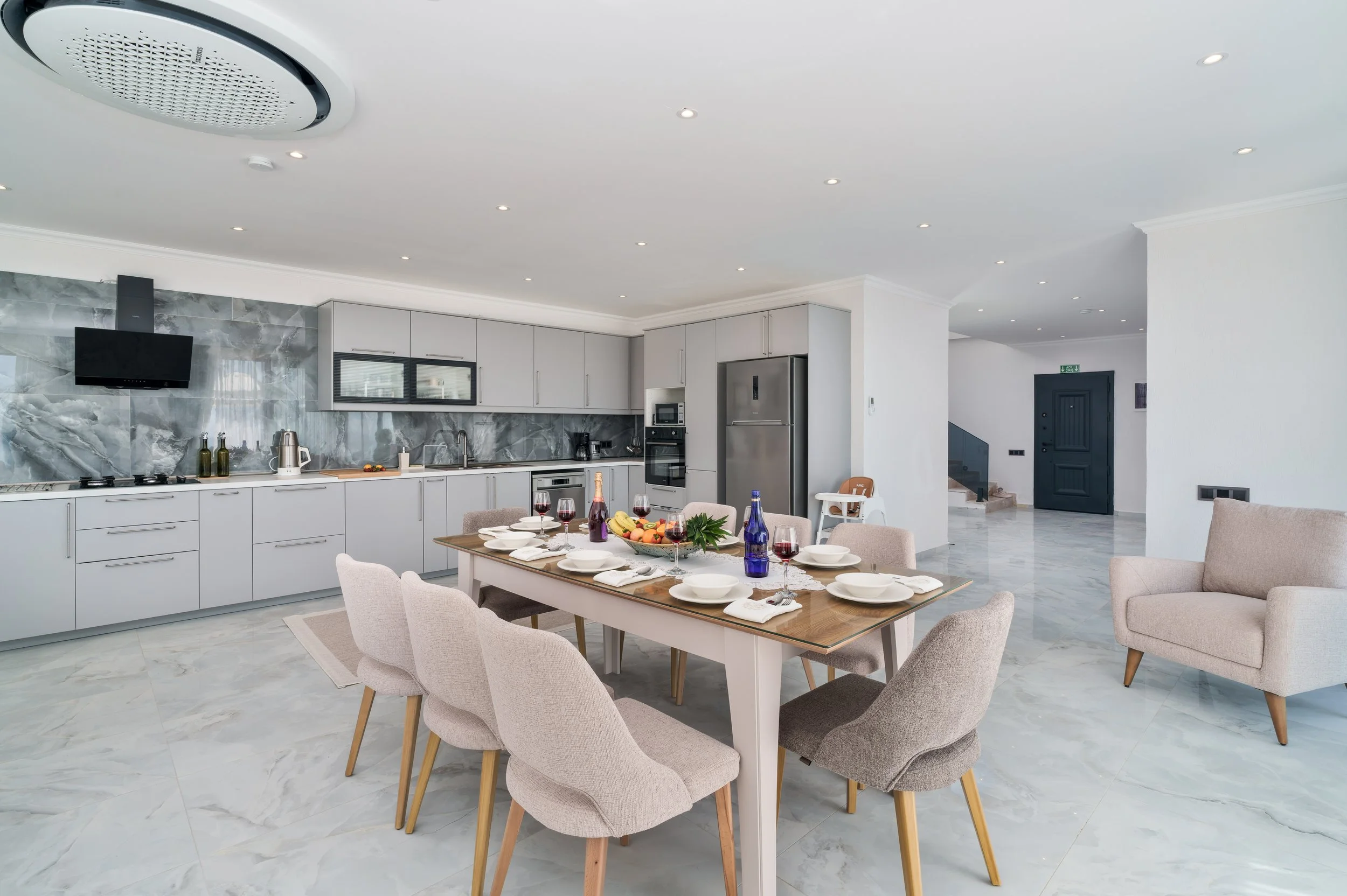 Modern kitchen with dining area, white cabinets, gray marble backsplash, and a dining table set with plates, wine glasses, and a bowl of fruit.