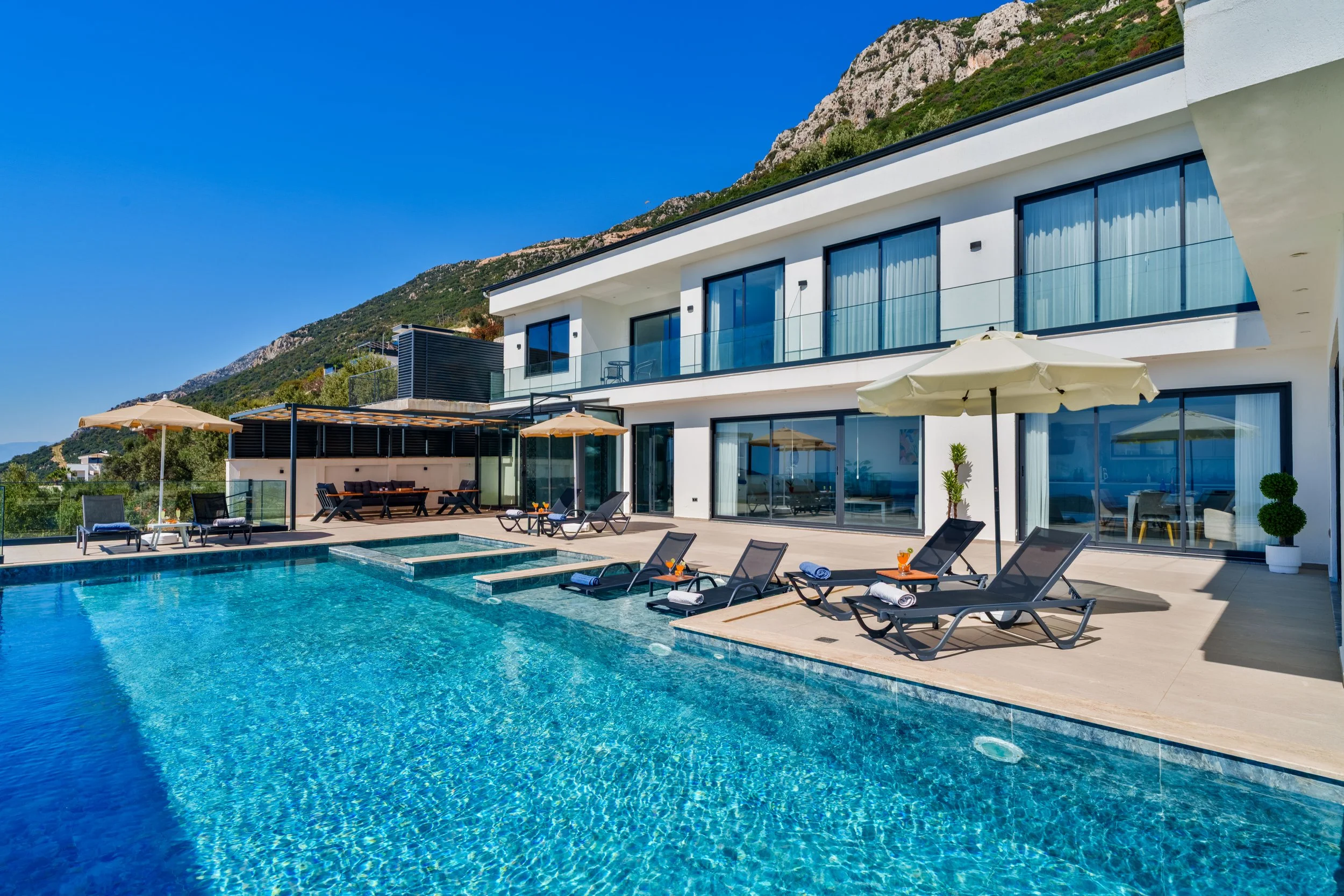 Modern white multi-story house with large glass doors and a swimming pool in the backyard, surrounded by lounge chairs and umbrellas, with a mountain in the background under a clear blue sky.