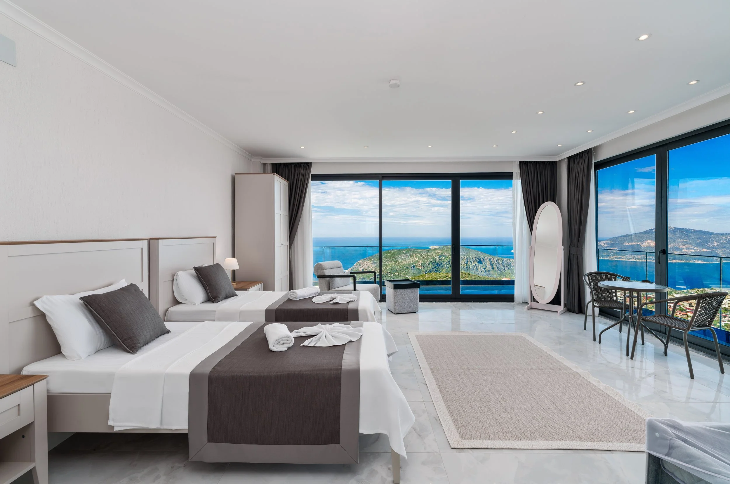 Spacious hotel room with two beds, large windows showing a scenic view of hills and the ocean, and modern furnishings.